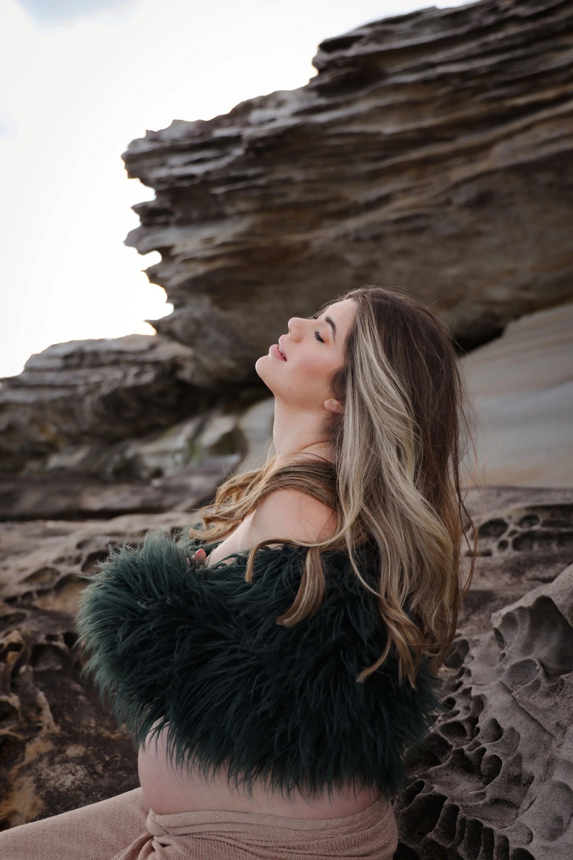 A woman with long, wavy hair and makeup, wearing a black fur stole and beige clothing, sitting on sand near large rock formations with an overcast sky.