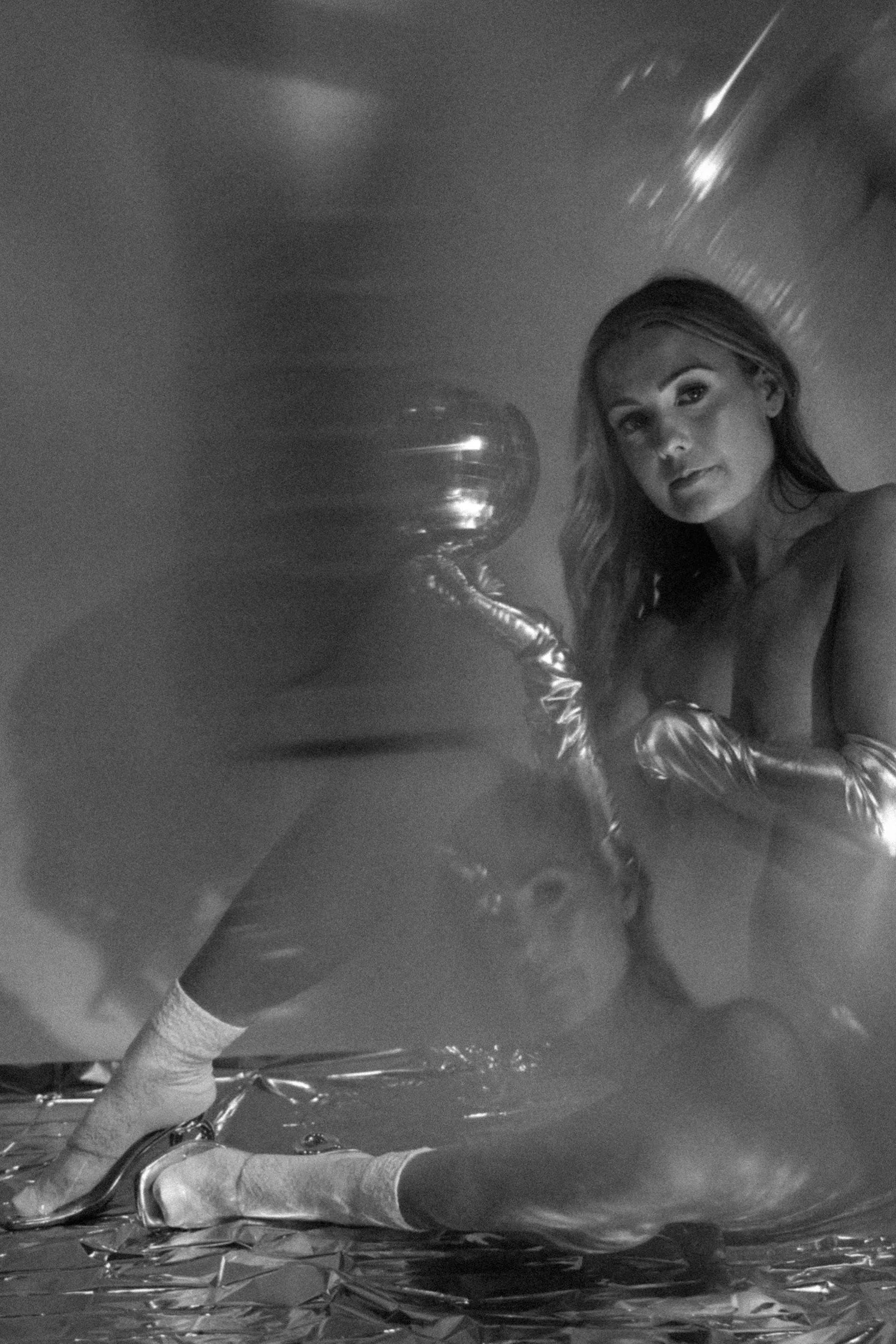 A woman with long hair and wearing shiny, metallic gloves is kneeling on a reflective surface, looking at the camera in a black and white photograph. The background appears to be a smooth, plain surface with some light reflections.