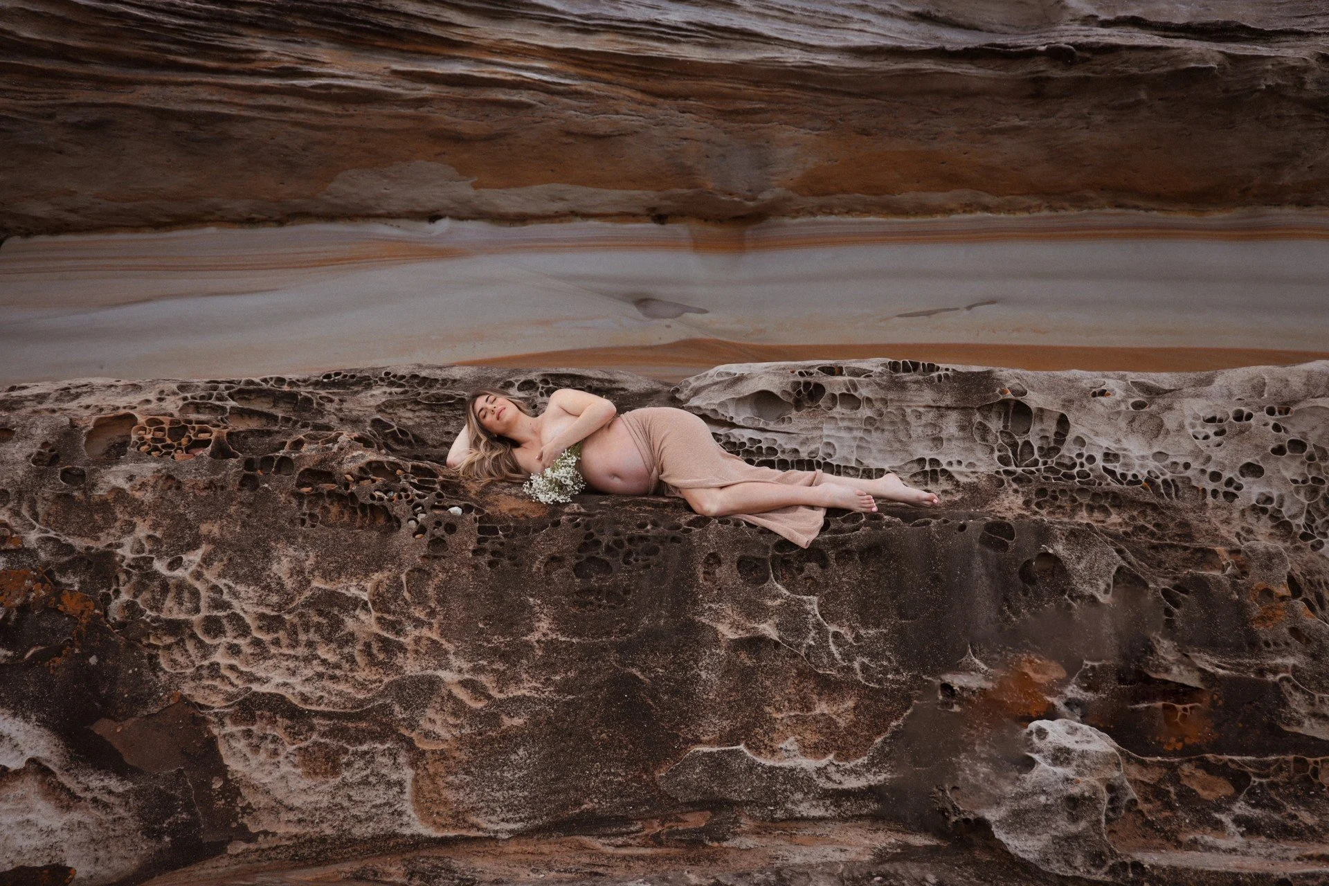A woman lying on a textured rock formation in a desert landscape, holding a small bouquet of white flowers.