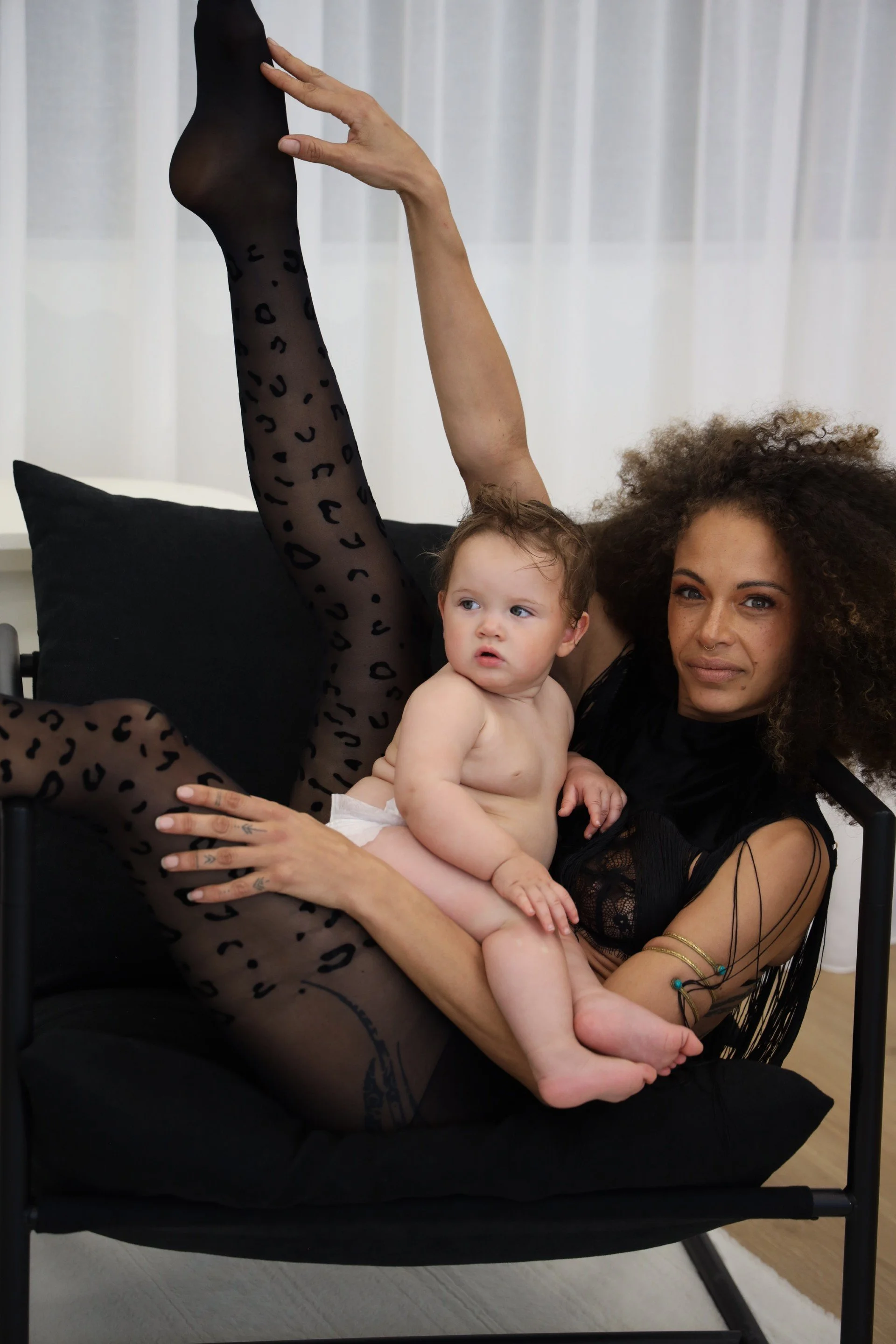 A woman with curly hair and a tattoo on her arm lies on a black chair, holding a young boy with curly hair who is shirtless and wearing a diaper. She is dressed in black fishnet stockings with leopard print and black clothing, extending one leg upwar