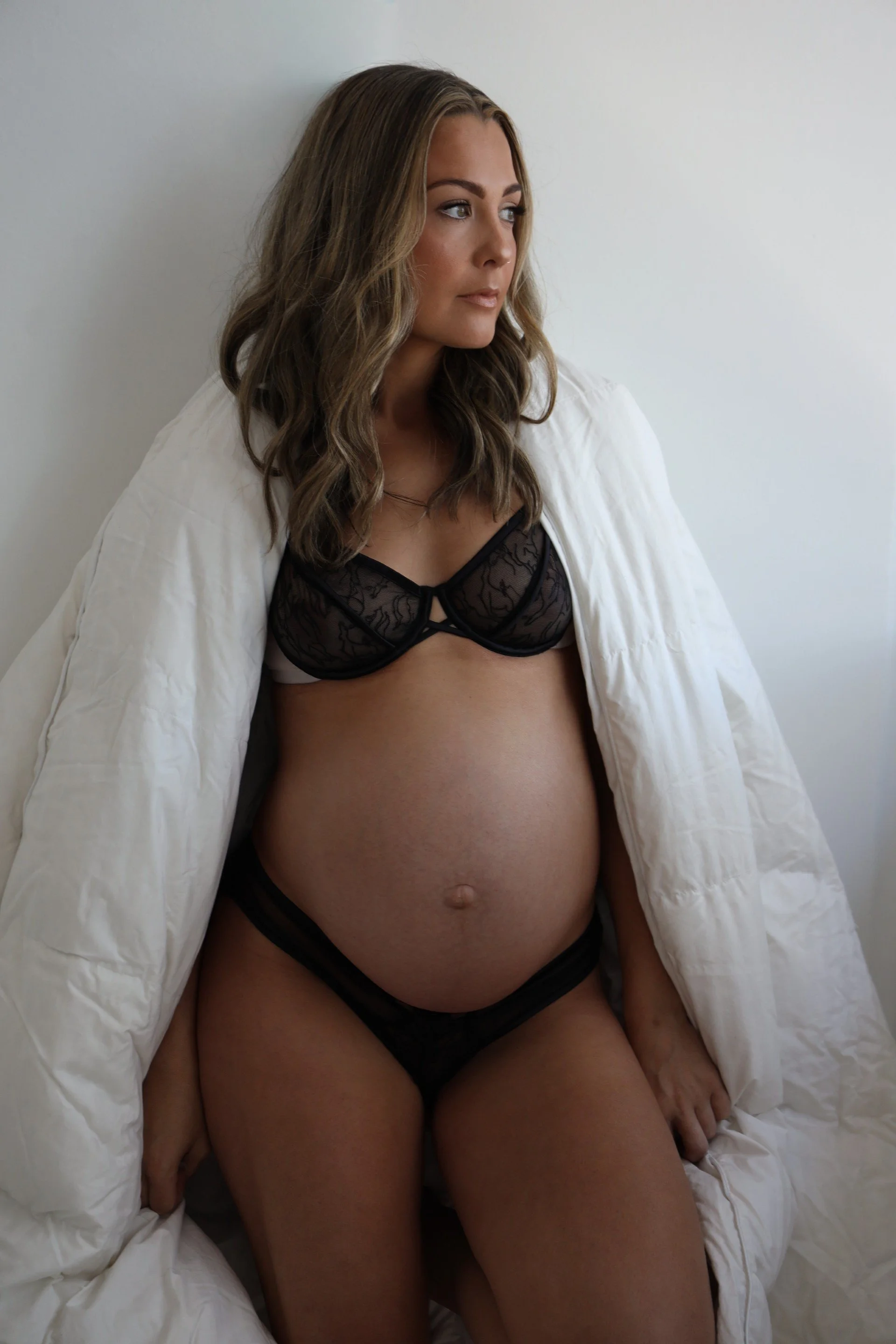 A pregnant woman with wavy brown hair wearing black lace lingerie and a white jacket over her shoulders, sitting against a plain white background.
