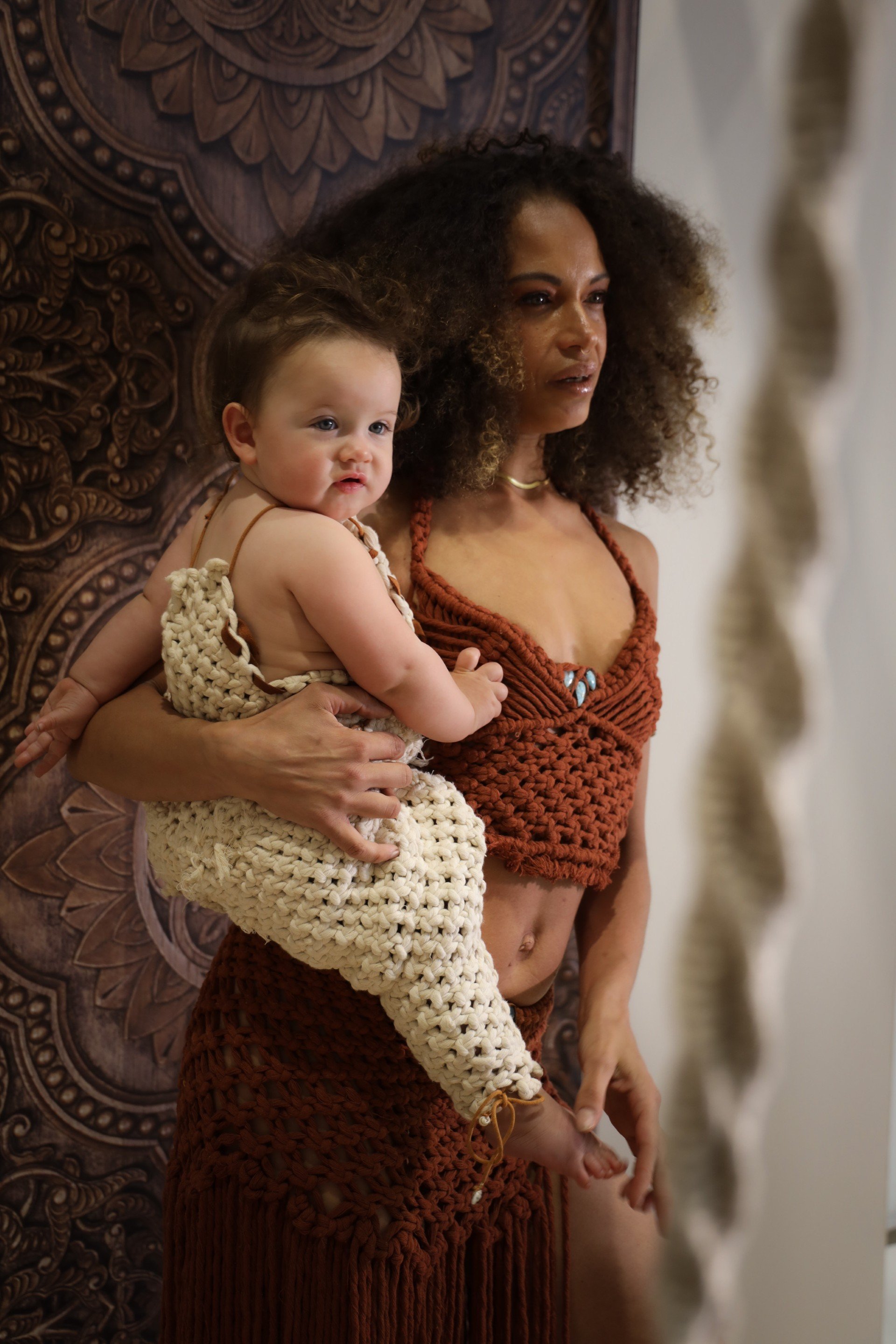 A woman holding a baby in her arms, standing next to a carved wooden panel and a stone sculpture.