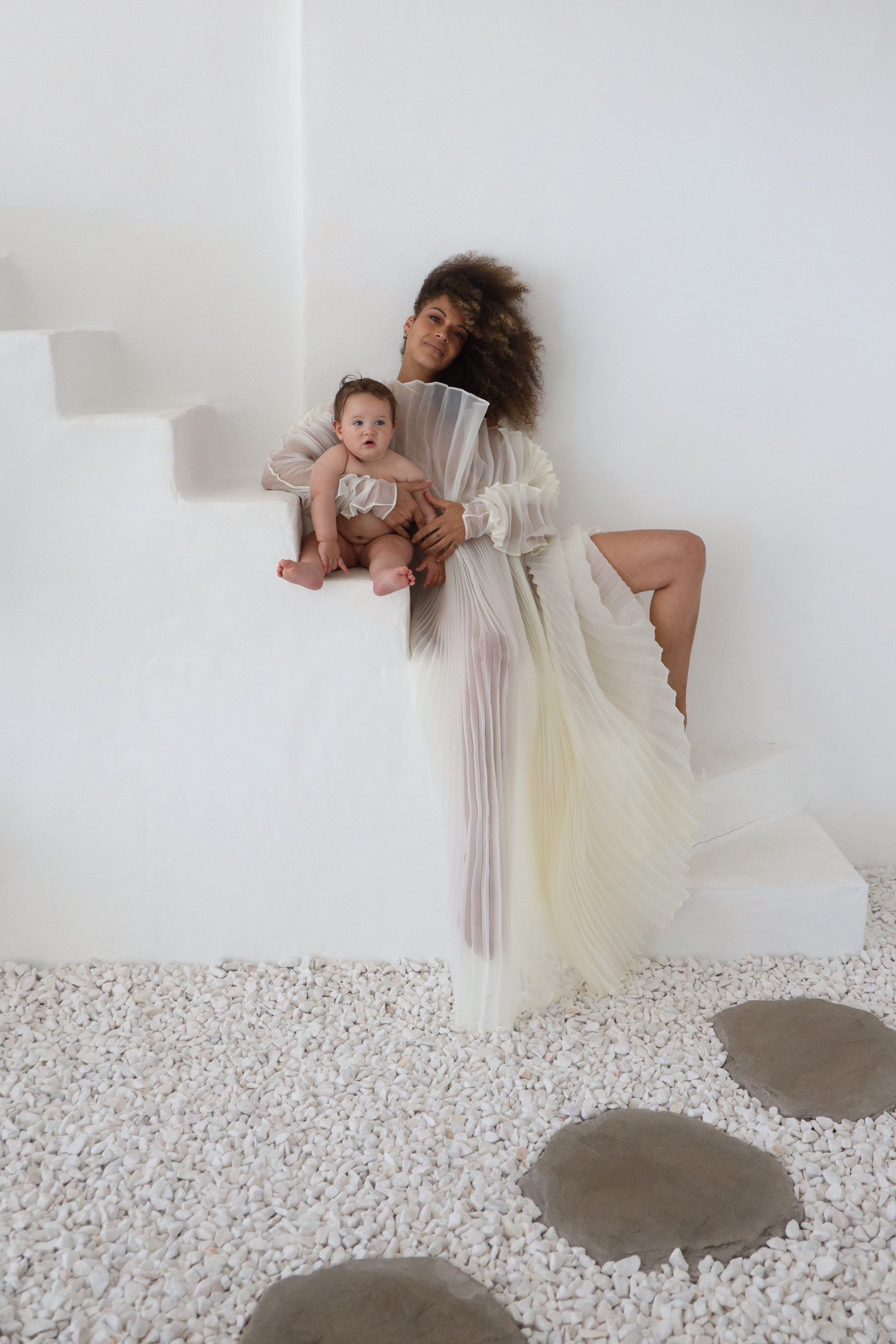 Woman dressed in a flowing, pleated white dress sitting on white steps with a baby resting on her lap, against a minimal white background with gravel and stepping stones on the ground.