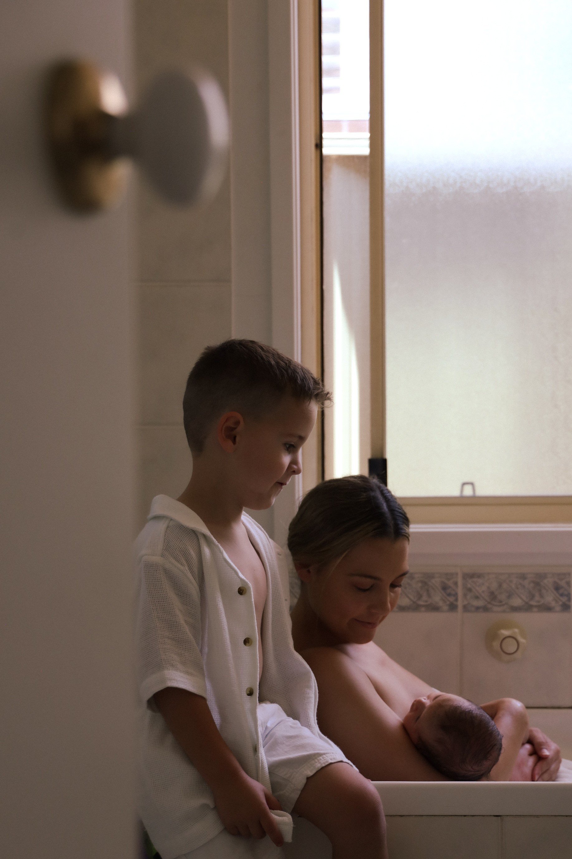 A woman holding a newborn baby in a bathtub, with a young boy sitting beside them in a bathroom.