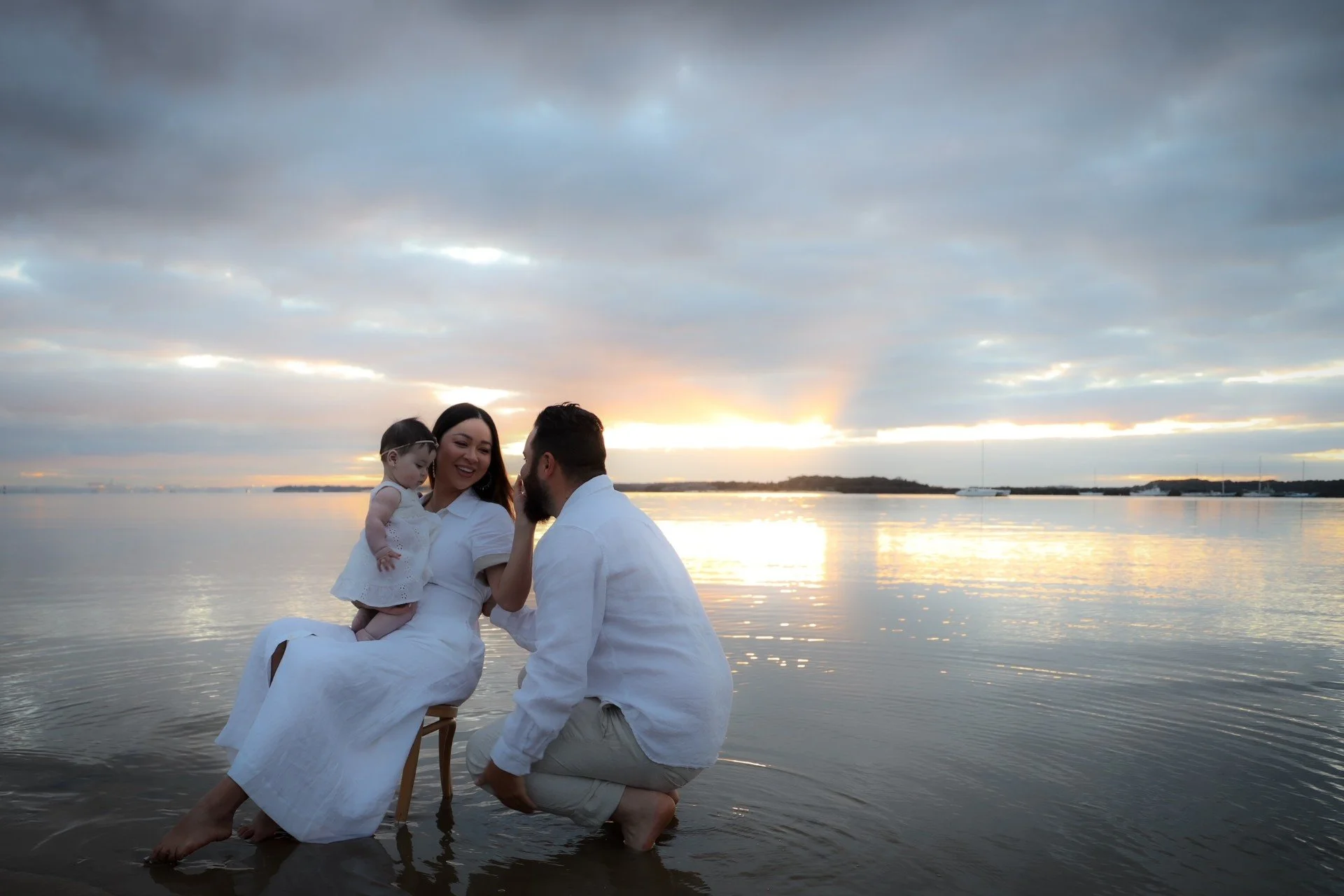 A family enjoying a moment by the water at sunset, with a woman holding a young girl on a chair, a man kneeling nearby, all dressed in white, by a calm body of water under a cloudy sky.