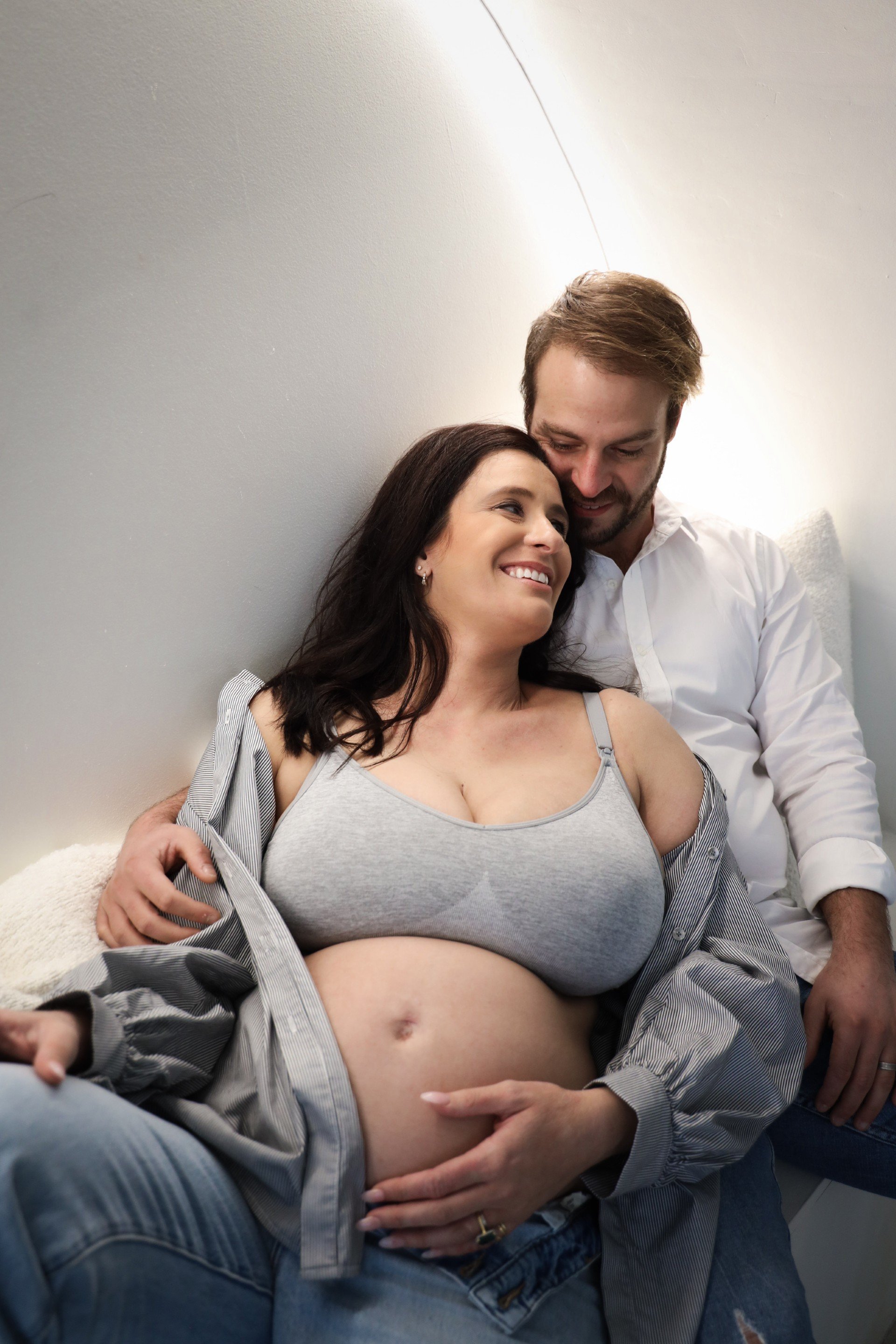 A pregnant woman and a man sitting closely together, the woman smiling and looking at the man, with the man's head resting near her forehead, in a cozy indoor setting.