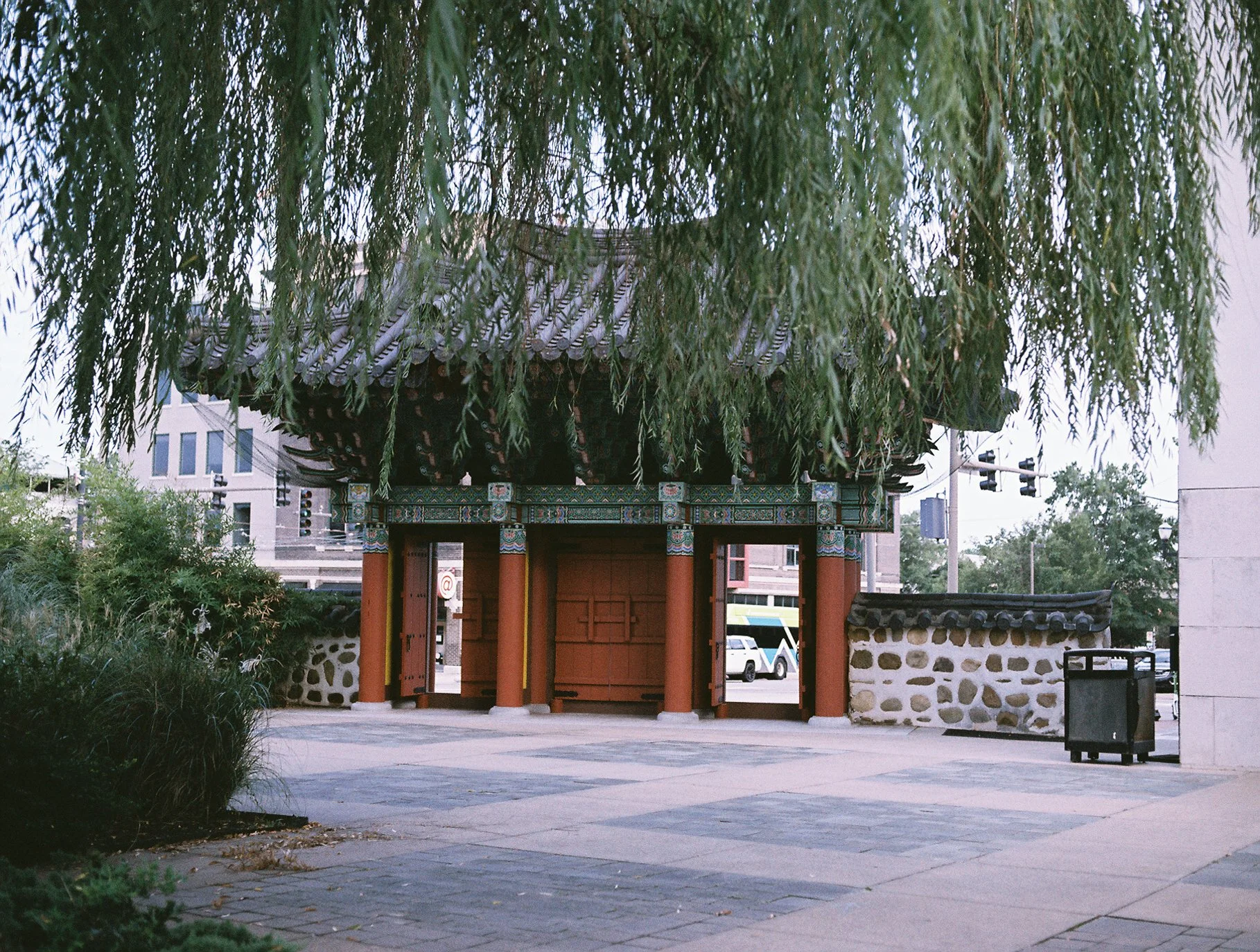 Ornate traditional gate with red columns, a curved tiled roof, and colorful painted details, framed by hanging willow branches.