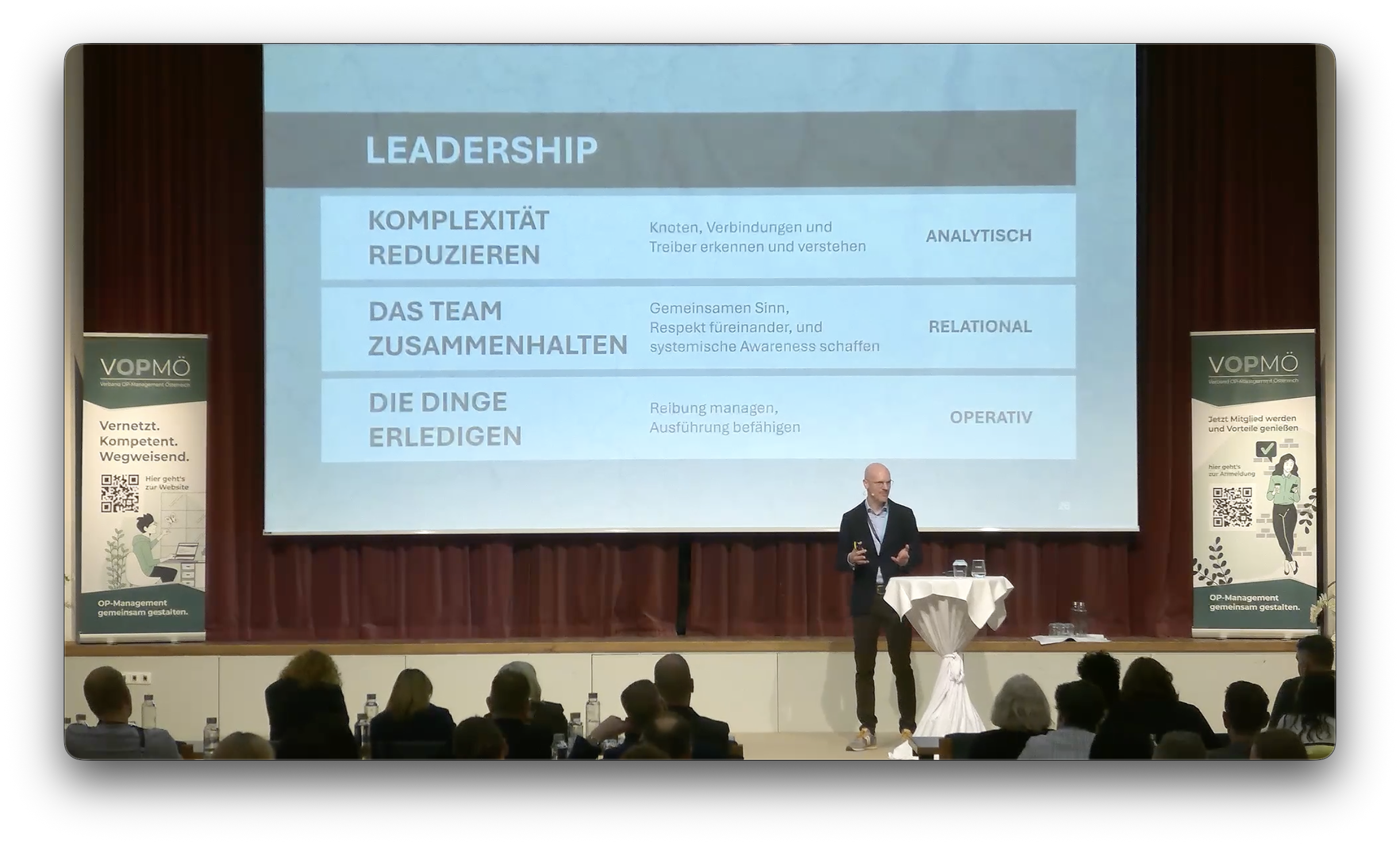 A man is giving a presentation on stage with a large screen behind him displaying a slide about leadership. The slide is titled 'Leadership' and contains text in German discussing complexity reduction, team cohesion, and task management with corresponding labels: analytical, relational, and operative. There are two vertical banners on either side of the stage, one featuring a QR code and a woman working on a laptop, and the other with a woman holding a microphone and a QR code. The audience is seated and watching the presentation.
