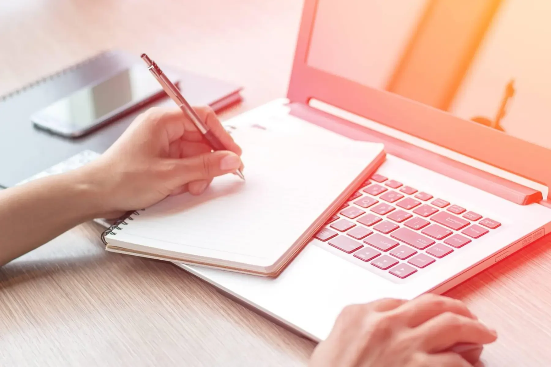 A pair of hands writing on a note book with a pen, laptop on a wooden table, orange tint to the image