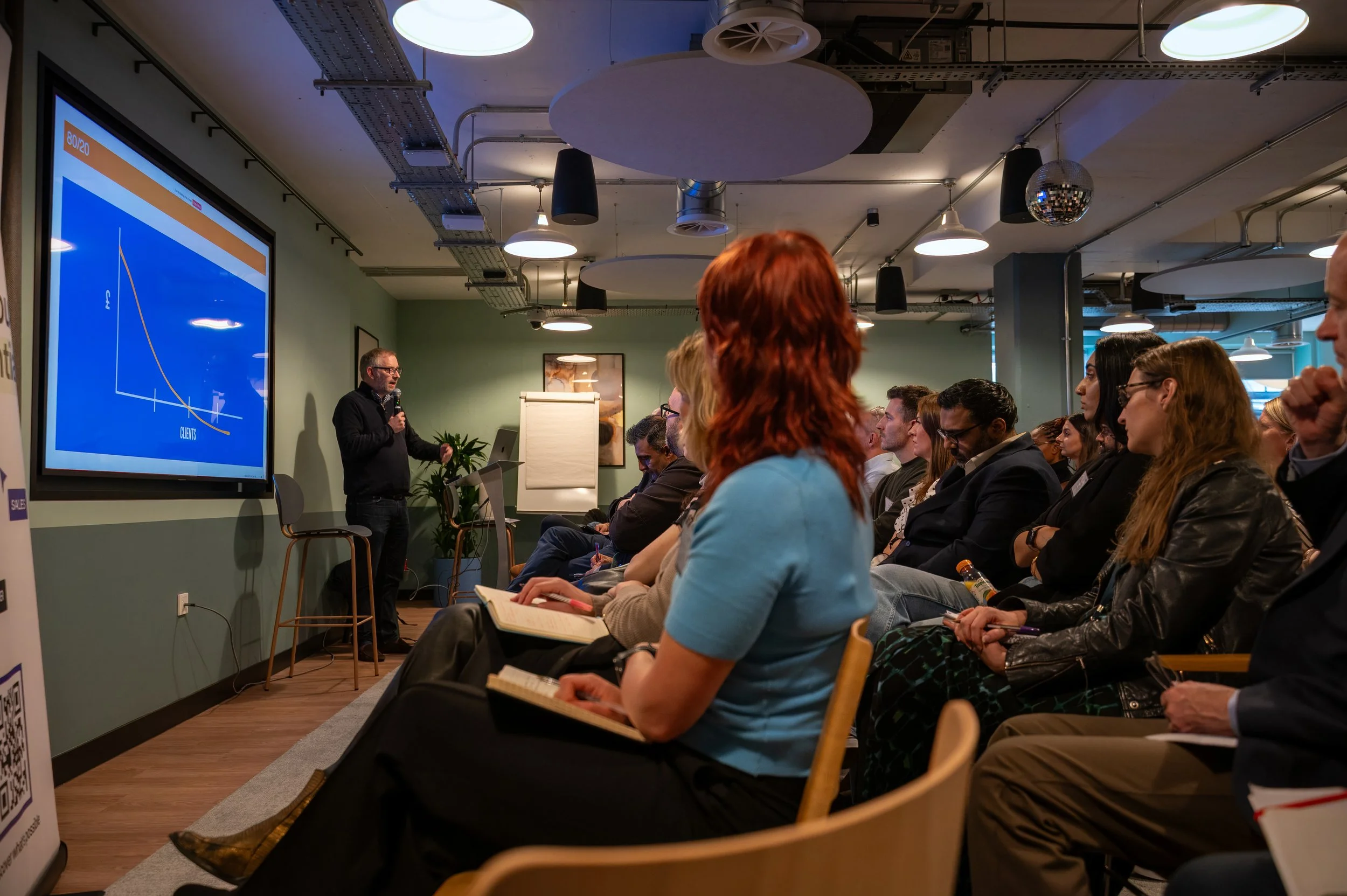 A professional workshop in a modern conference room, where Jeremy Brim, the speaker, presents a slide featuring a graph. Attendees are seated, engaged, and taking notes in a casual business setting.