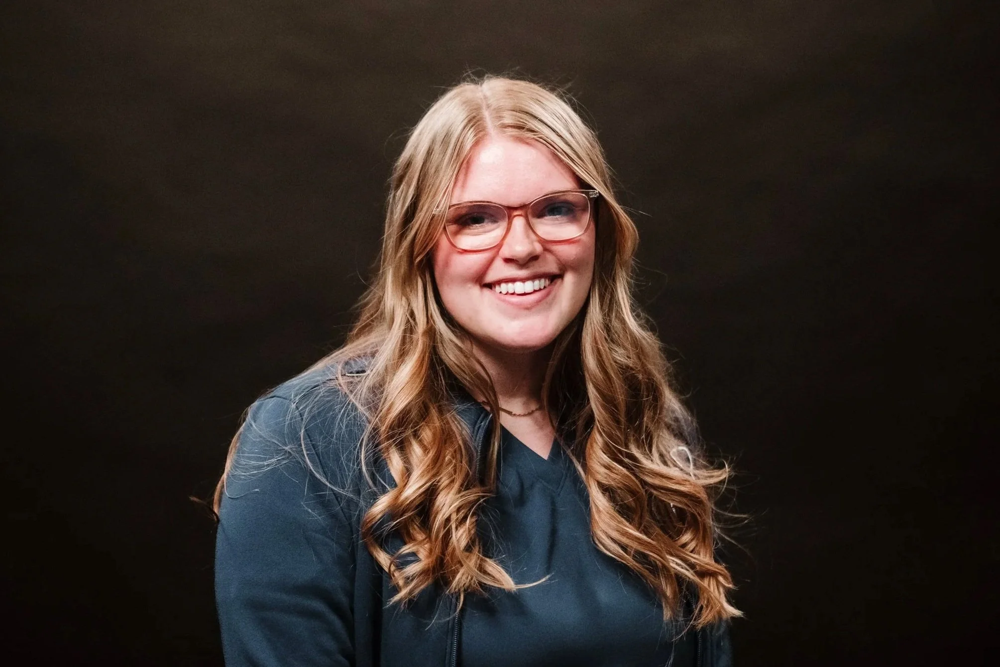 A woman with long wavy light brown hair, wearing glasses and a dark blue top, smiling against a dark background.