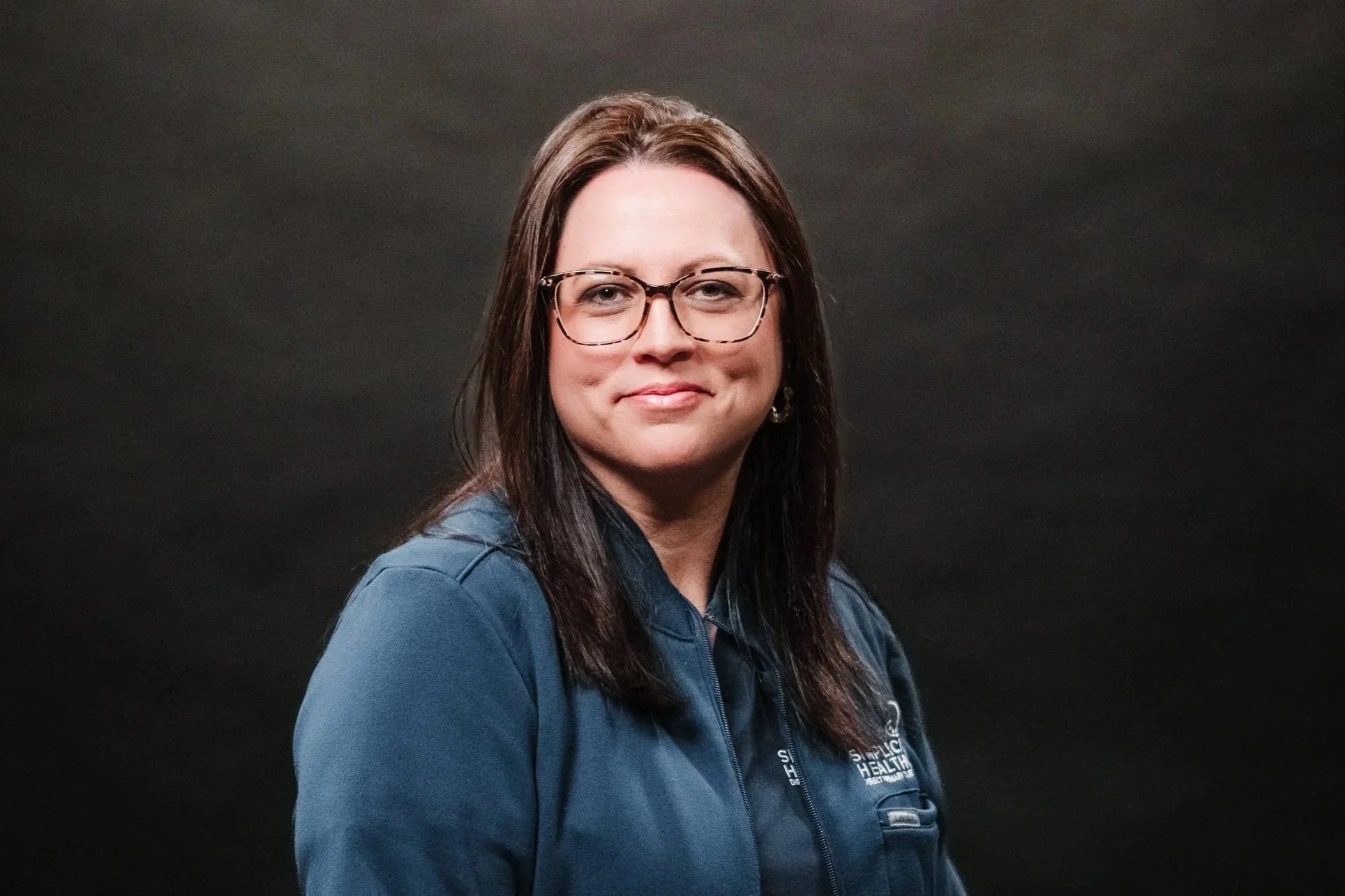 A woman with dark brown hair, glasses, and light makeup, wearing a blue jacket, is smiling slightly against a dark gray background.