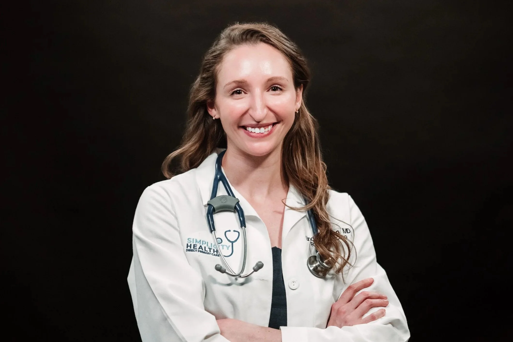 A smiling, Dr. Marton, a female doctor with long, wavy brown hair wearing a white coat with a stethoscope around her neck, standing against a dark background.