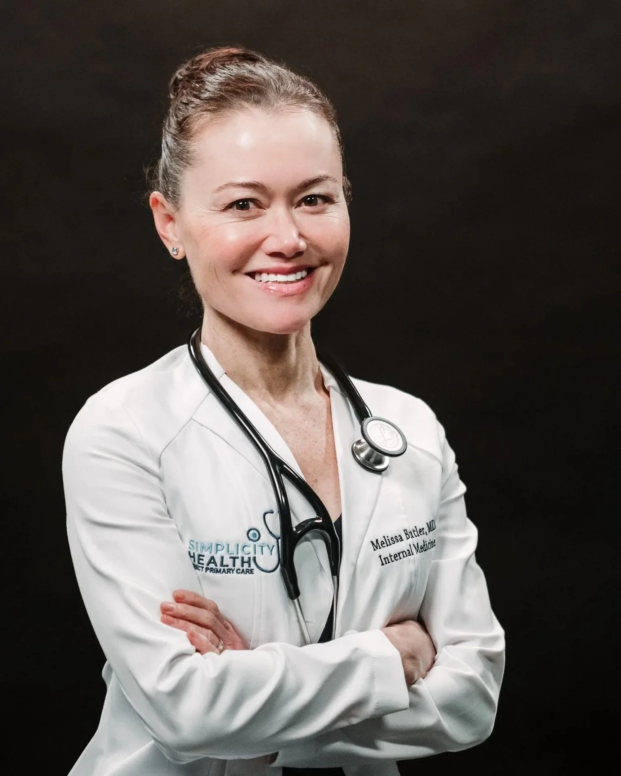 A smiling, Dr. Butler, a female doctor wearing a white coat with a stethoscope around her neck, standing against a dark background.