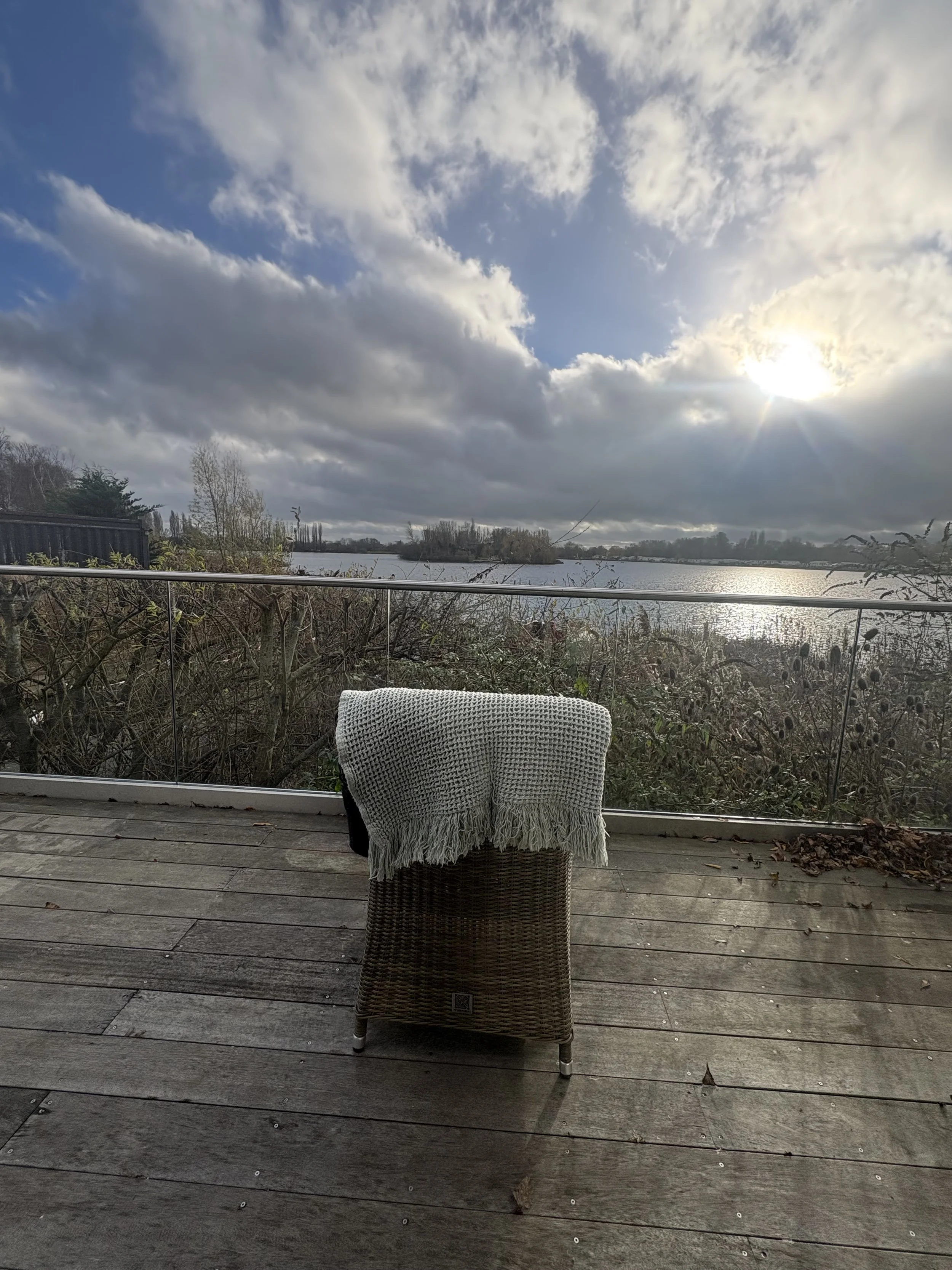 A wicker chair with a white knitted blanket draped over it on a wooden deck overlooking a lake, under a cloudy sky with the sun partially visible.