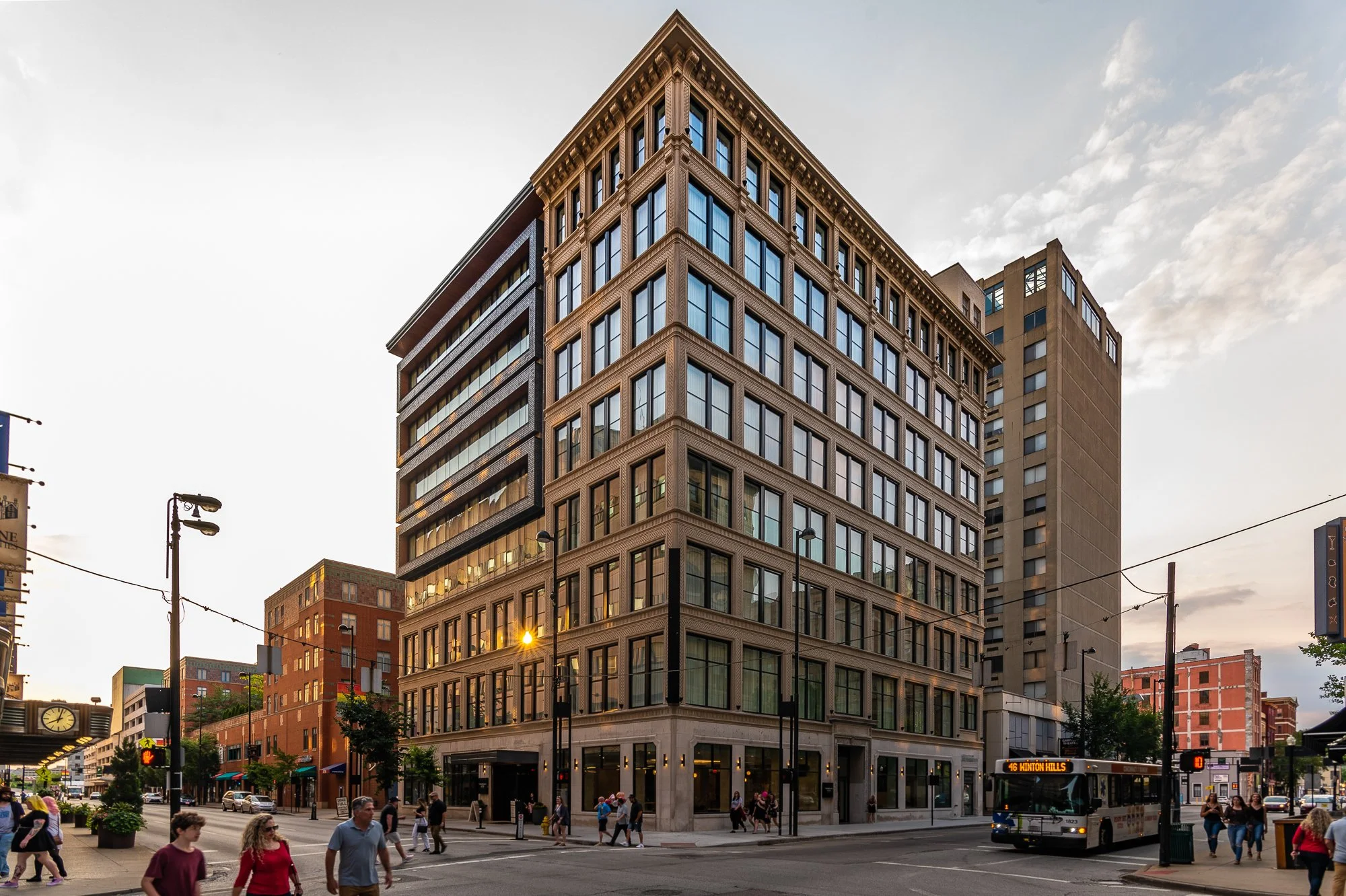 A multi-story brick building with large windows on street corner, with pedestrians, a bus, and streetlights in an urban setting at dusk.