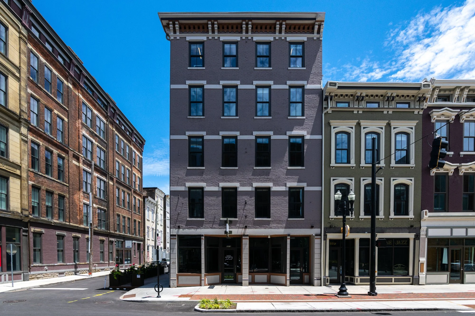 Four-story brick building on city street with mixed-use ground floor, surrounded by other historic style buildings, under a blue sky with some clouds.