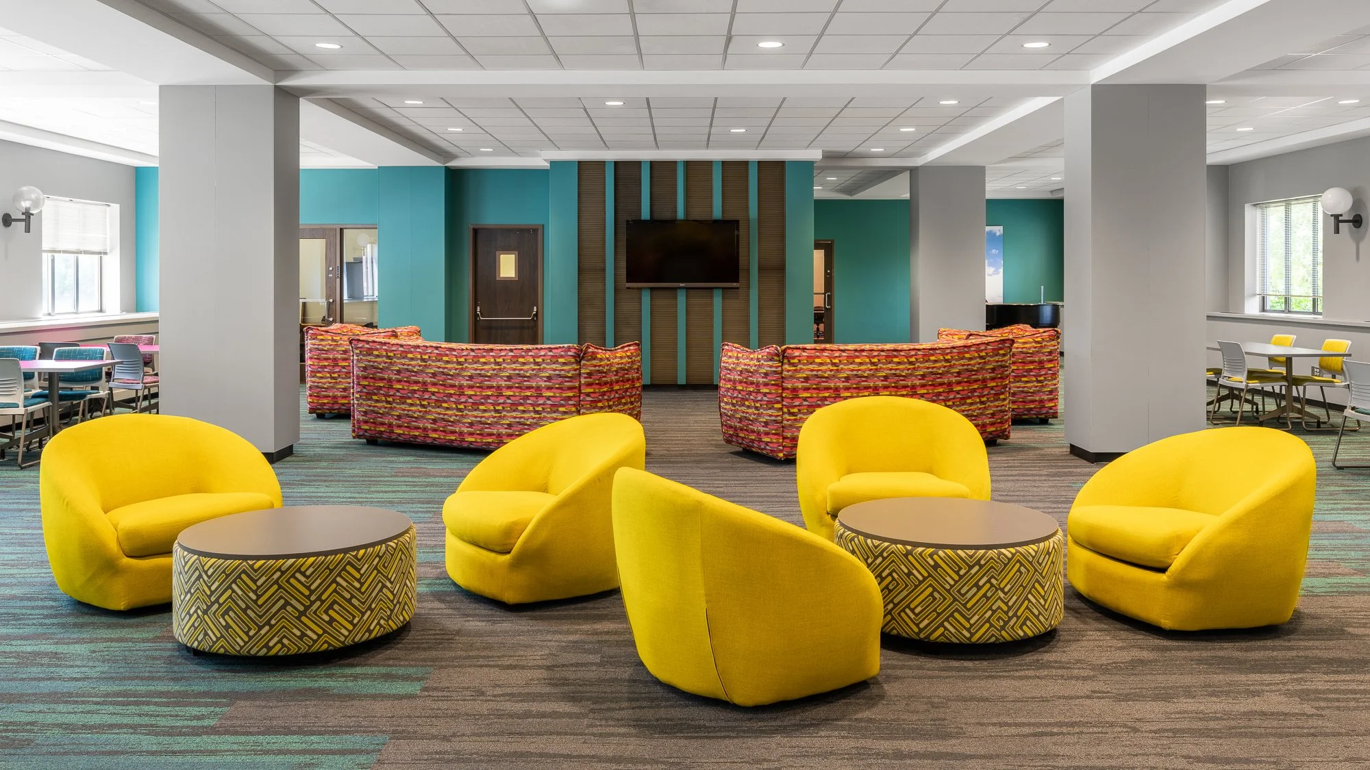 Colorful lobby with yellow chairs and patterned ottomans, teal accent wall, and a television on a wooden panel amid seating areas.
