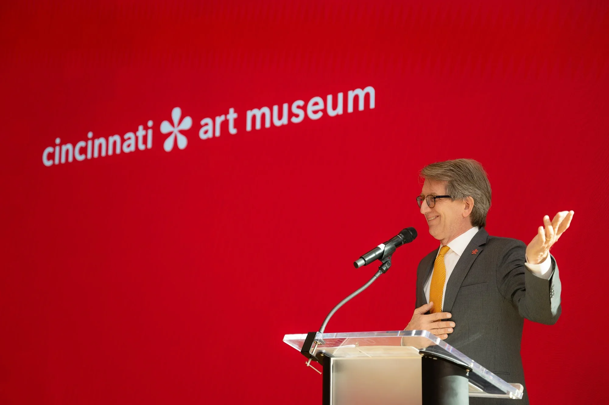 A man in a suit and yellow tie standing at a podium with a microphone, smiling and gesturing with one hand, in front of a red background with the Cincinnati Art Museum logo.