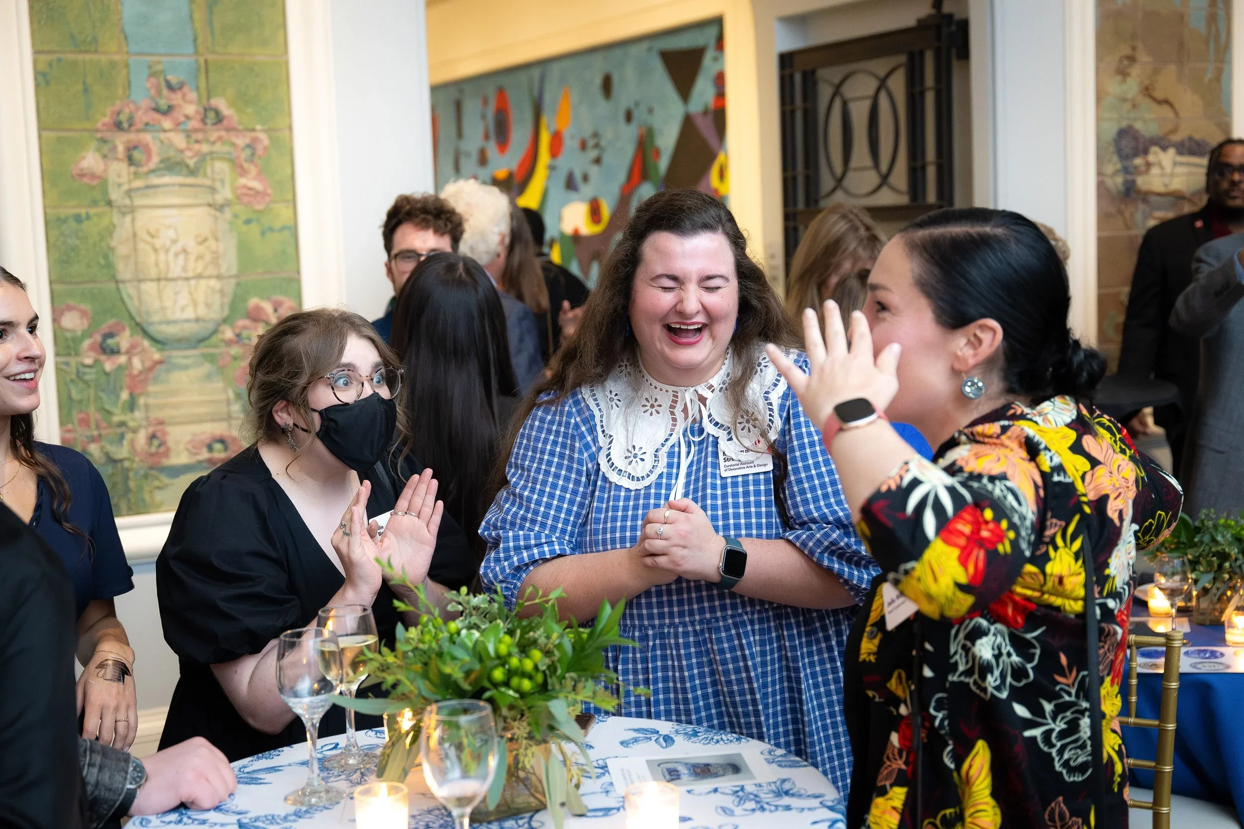 Group of women at a social gathering, smiling and laughing while standing around a table with a floral centerpiece, glasses, and lit candles, indoors with colorful abstract art on the wall.