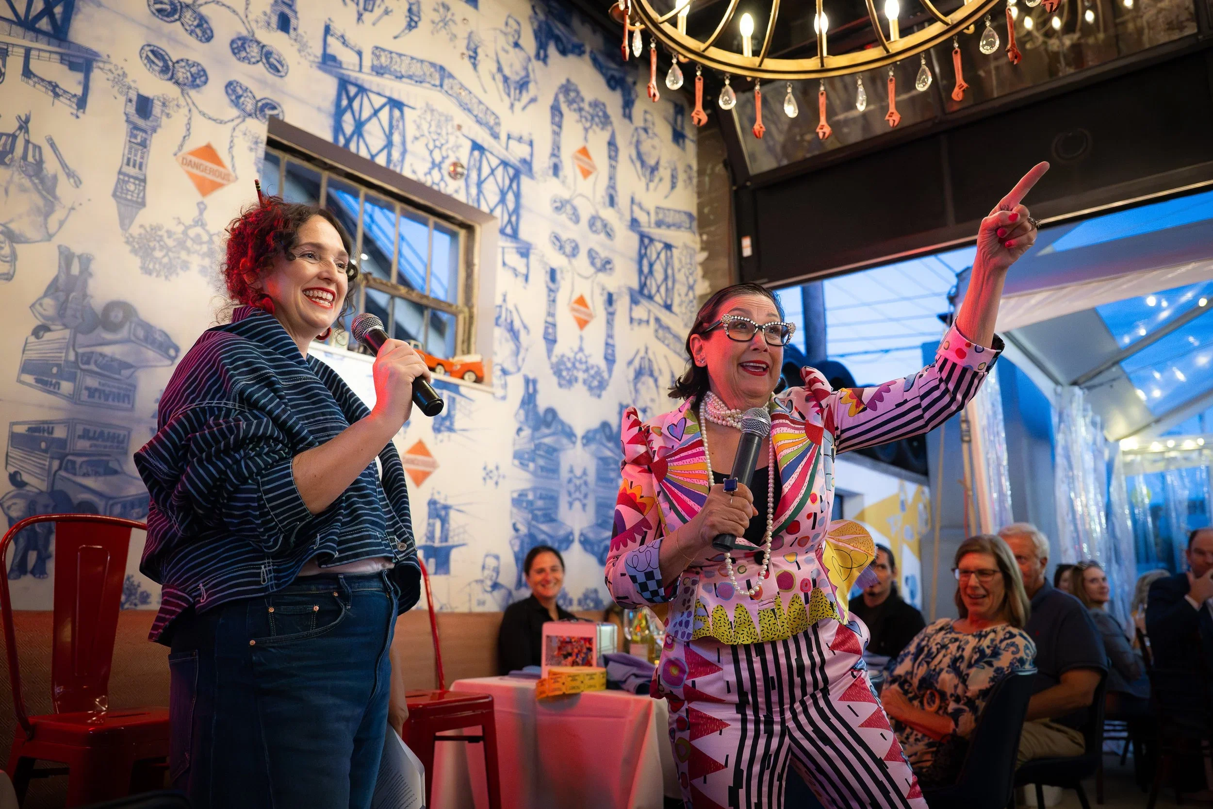 Two women standing on a stage, holding microphones, speaking to an audience; lively atmosphere with people seated at tables, decorated with colorful artwork and murals, in an artsy indoor venue.