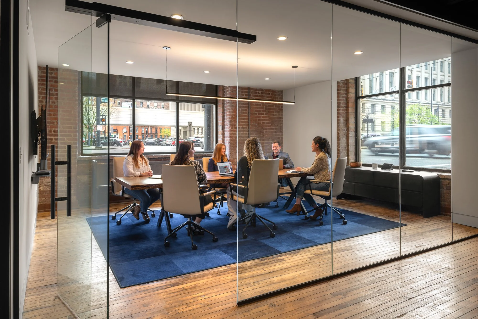 A meeting room with glass walls, wooden floors, a large wooden table, and six women and one man sitting around it, having a discussion during daytime.
