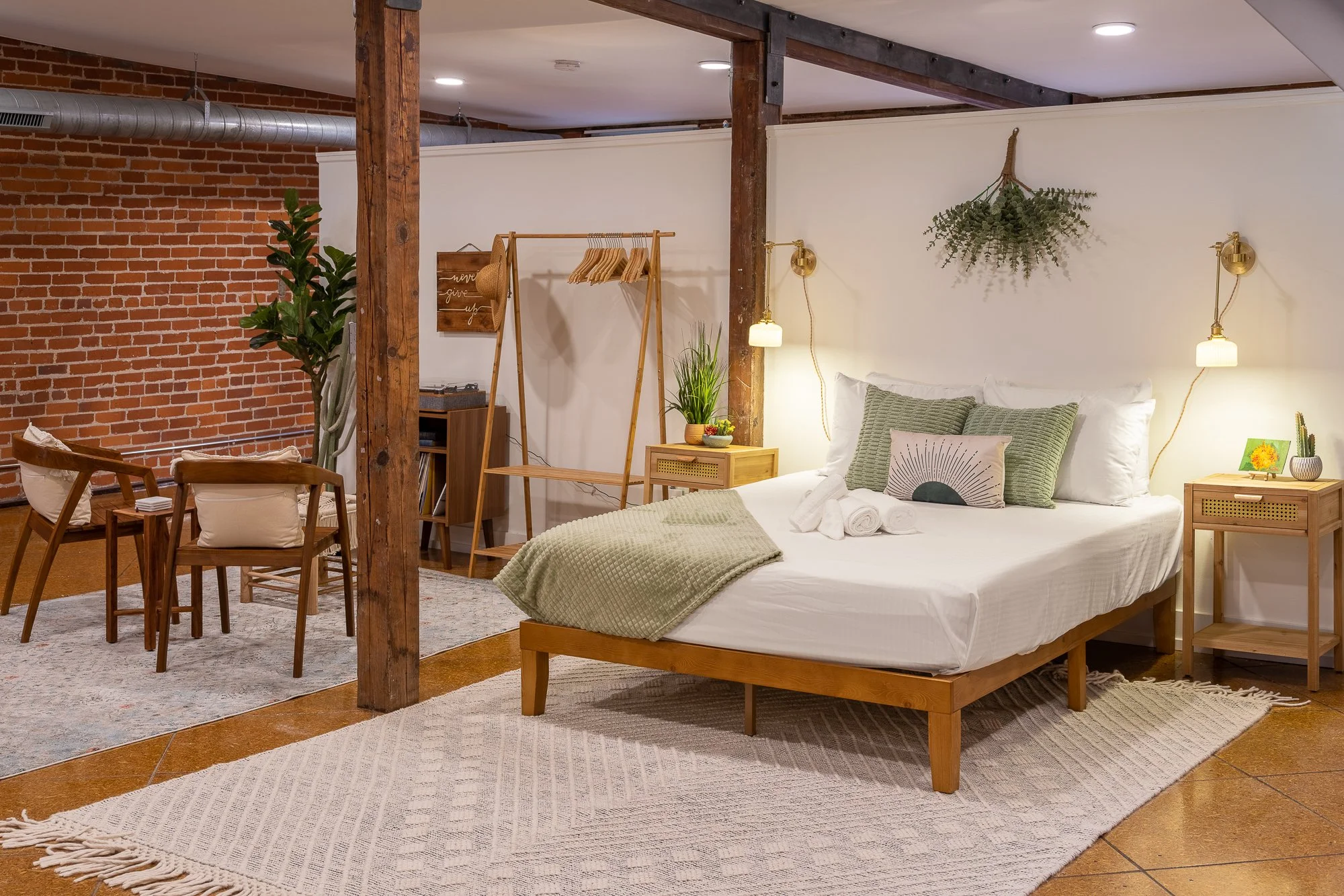 Cozy bedroom with a white bed featuring green and white pillows, a textured green blanket, and rolled towels. Wooden nightstands with lamps, a potted plant, and decorative wall hanging above the bed. Nearby, a wooden clothing rack, plant, and seating