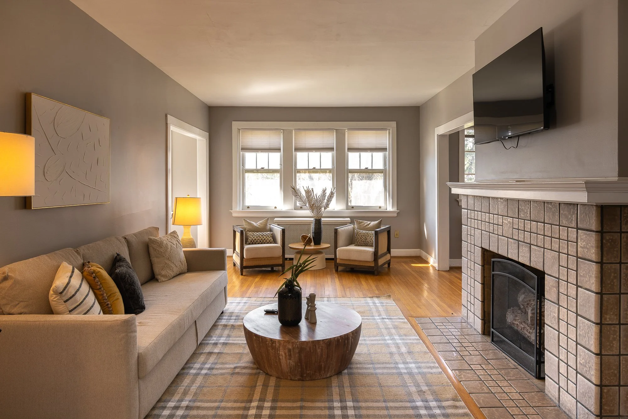 Living room with beige sofa, patterned pillows, round wooden coffee table, checkered rug, fireplace, wall-mounted TV, three corner armchairs near windows, side lamps, and decorative vases.