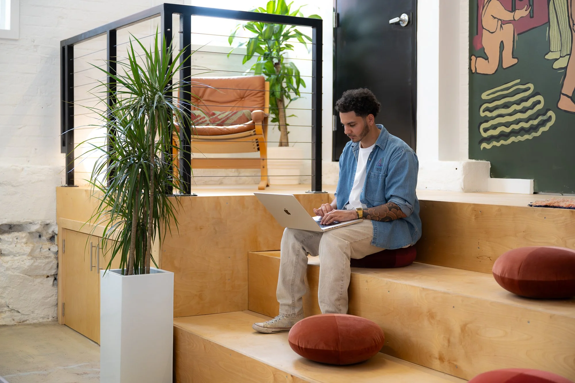 A man sitting on a wooden bench working on a laptop in a cozy, modern space with plants and a black chalkboard wall.