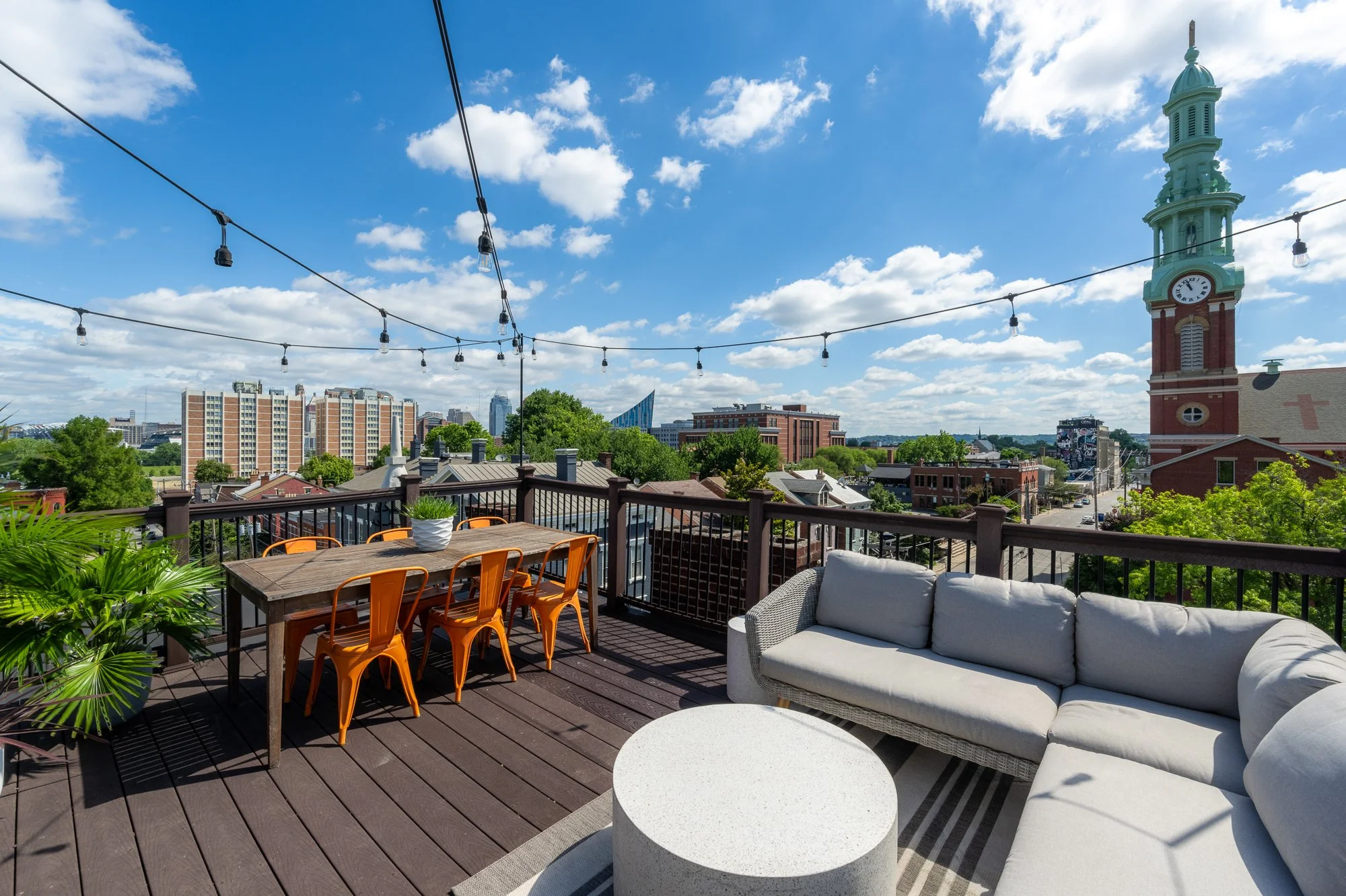 Rooftop patio with outdoor furniture including a gray couch, a round white table, and a wooden table with orange chairs. String lights overhead, city skyline with buildings and a church with a clock tower in the background, and a partly cloudy sky.