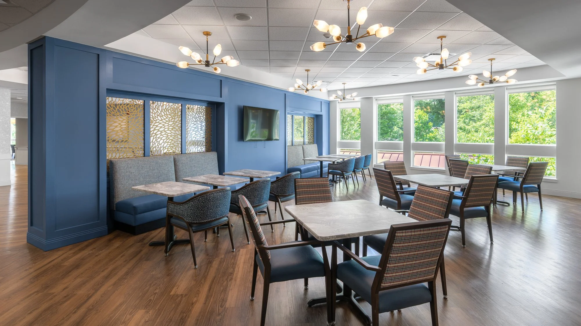 Empty dining area with tables and chairs, large windows with greenery outside, wooden flooring, blue accent wall, and modern chandelier lighting.