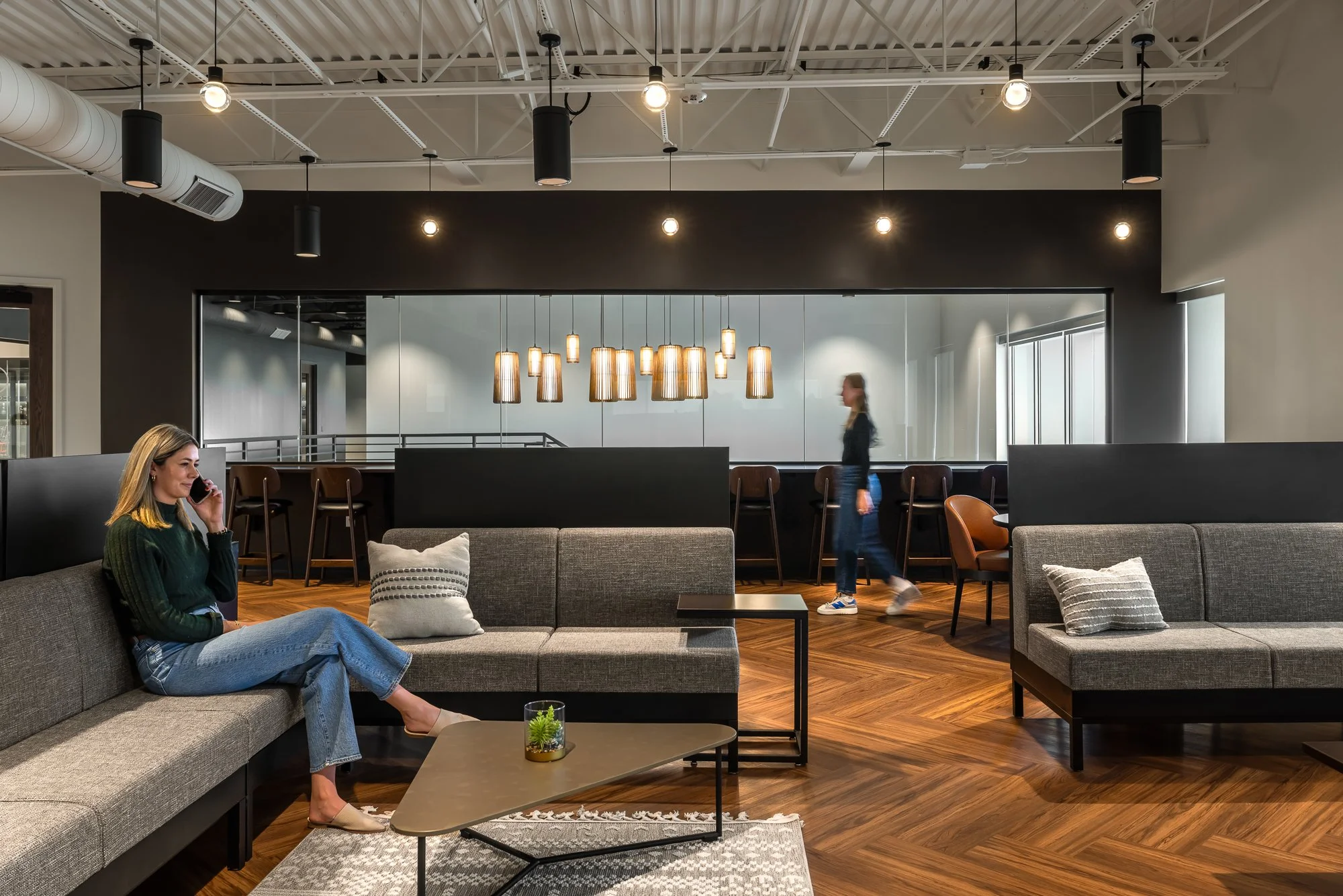 Modern lobby area with gray sofas, a wooden coffee table with a plant, a woman sitting and talking on her phone, and another woman walking past in the background.