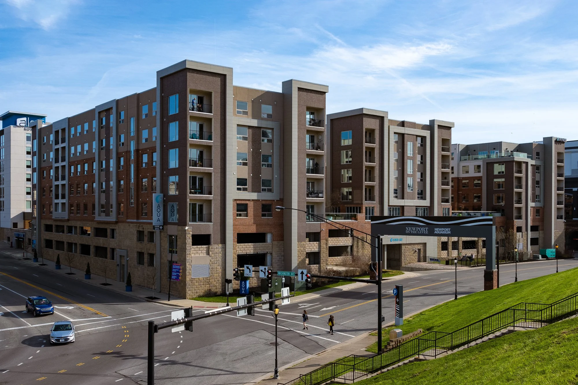 A multi-story residential building with brick and beige exterior, glass balconies, and various windows, located along a city street with cars, pedestrians, traffic signals, and street signs.
