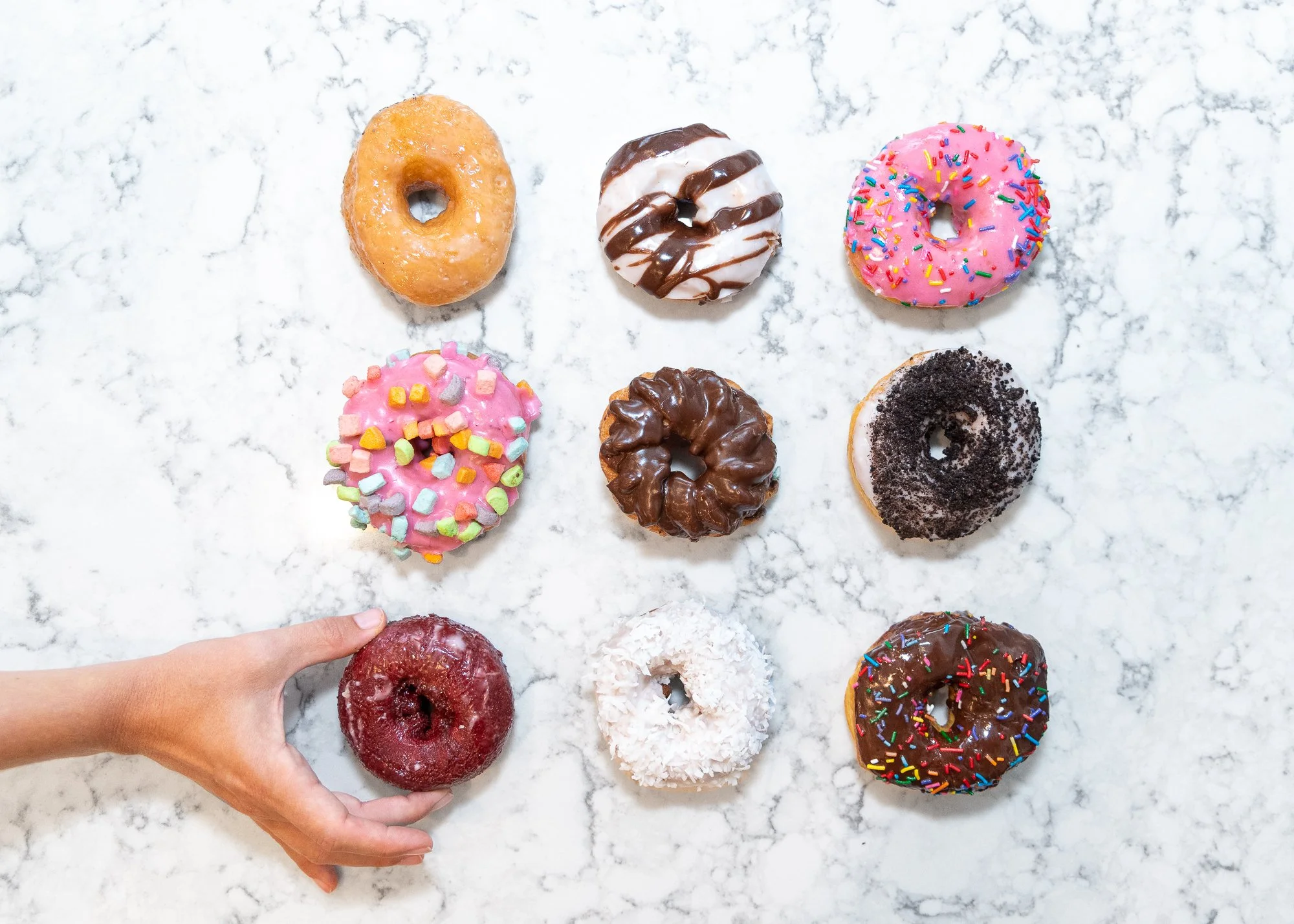 A hand picking up a red-glazed donut with sprinkling on a white marble surface. Surrounding it are eight other donuts with various toppings and flavors, arranged in three rows.