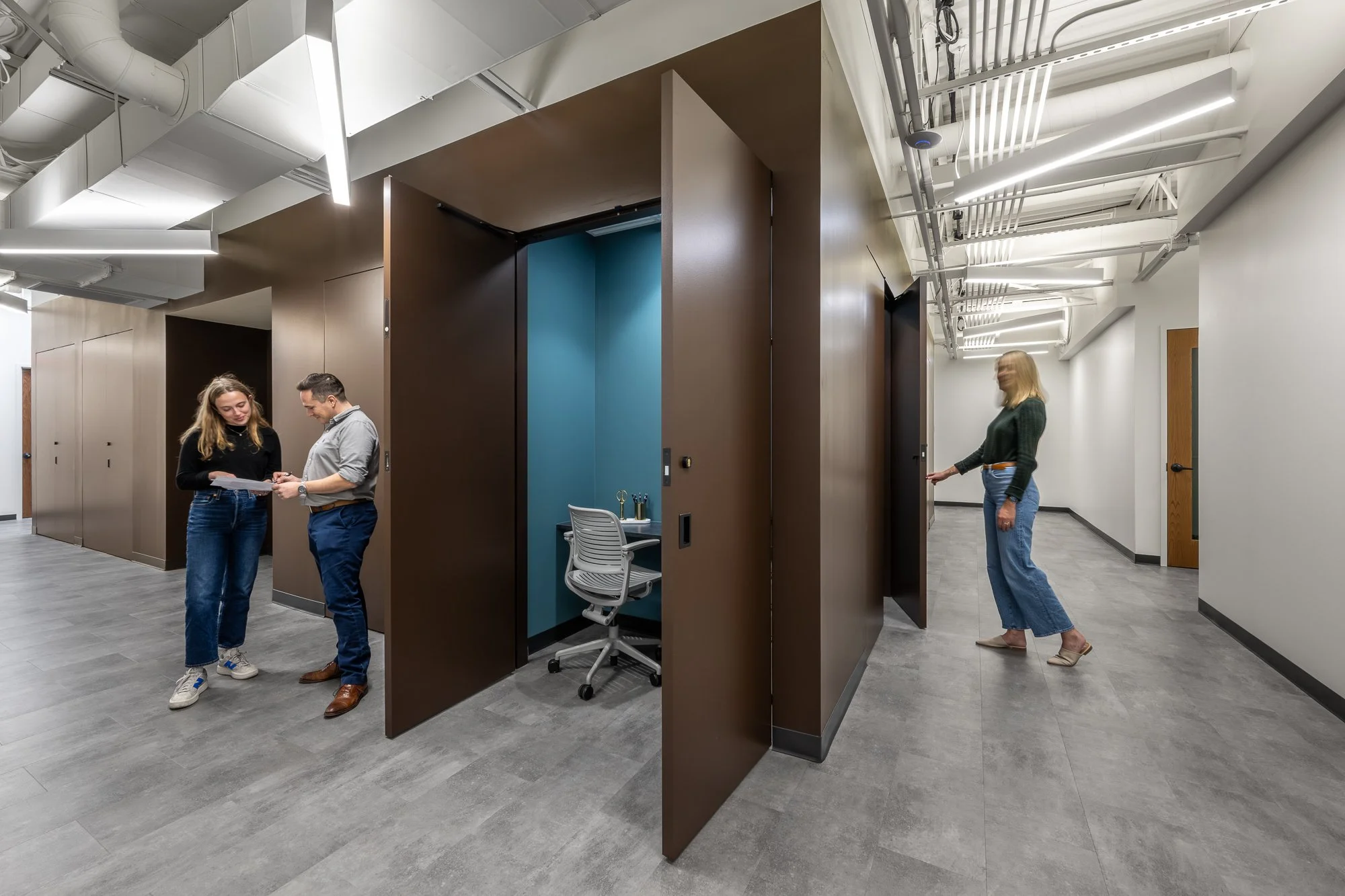 Two women and one man in an office hallway, with two small office booths and modern ceiling lights.