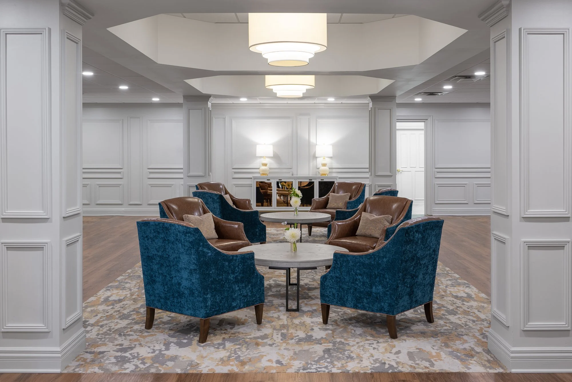 Elegant hotel lobby with six blue velvet and brown leather armchairs arranged around three small round tables, floral centerpieces, white paneled walls, and modern lighting fixtures.