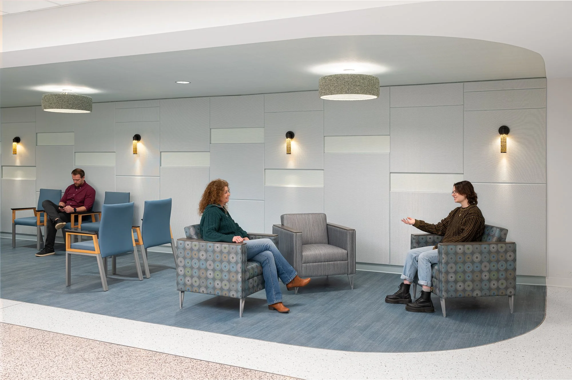 Three people sitting in a waiting area, two women and one man, with the women engaging in conversation and the man looking at his phone.