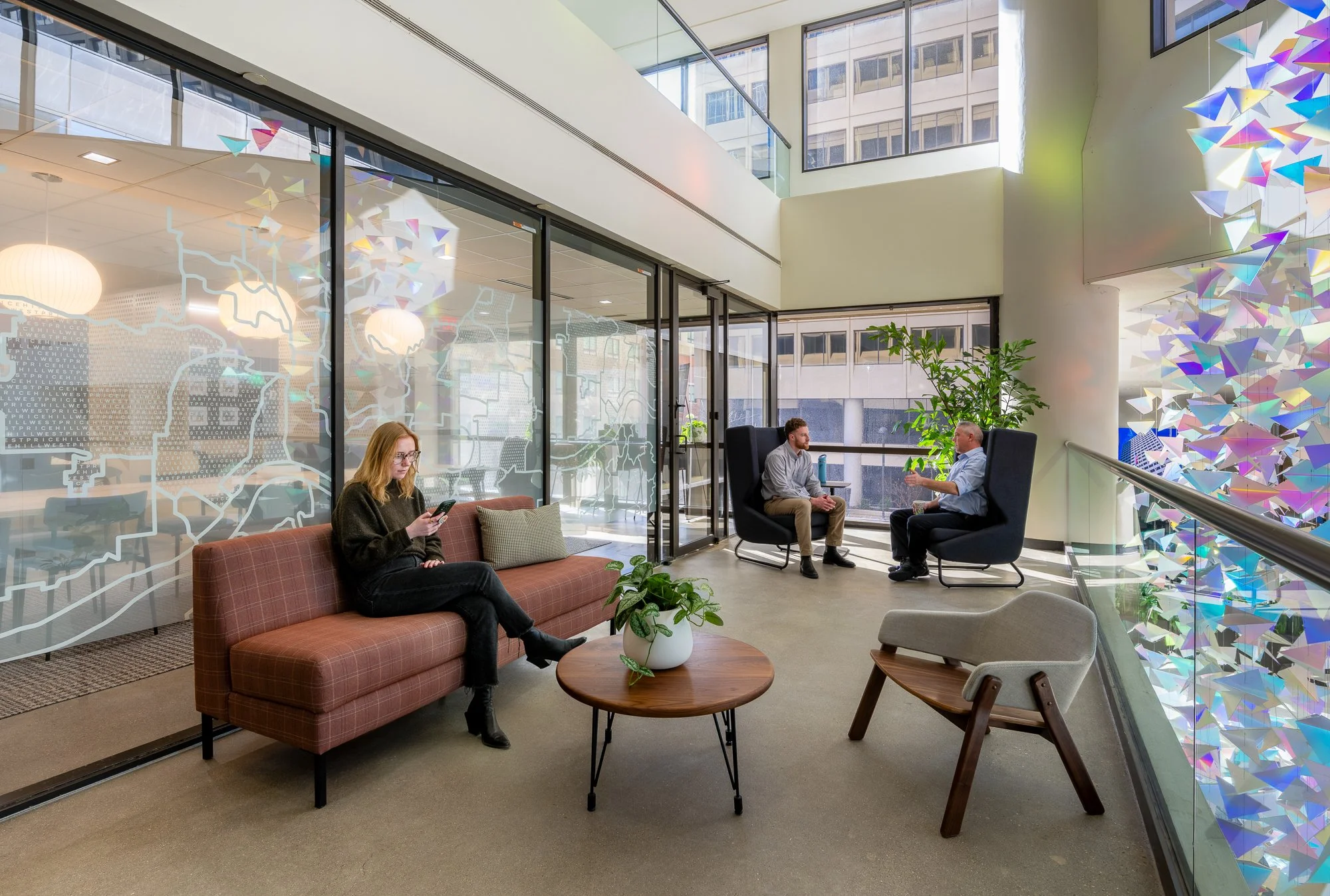 Interior view of a modern office lounge with glass walls, a brown sofa, a woman using her phone, two men sitting in high-backed chairs having a conversation, a small round table with a plant, and colorful decorative hanging art on the wall.