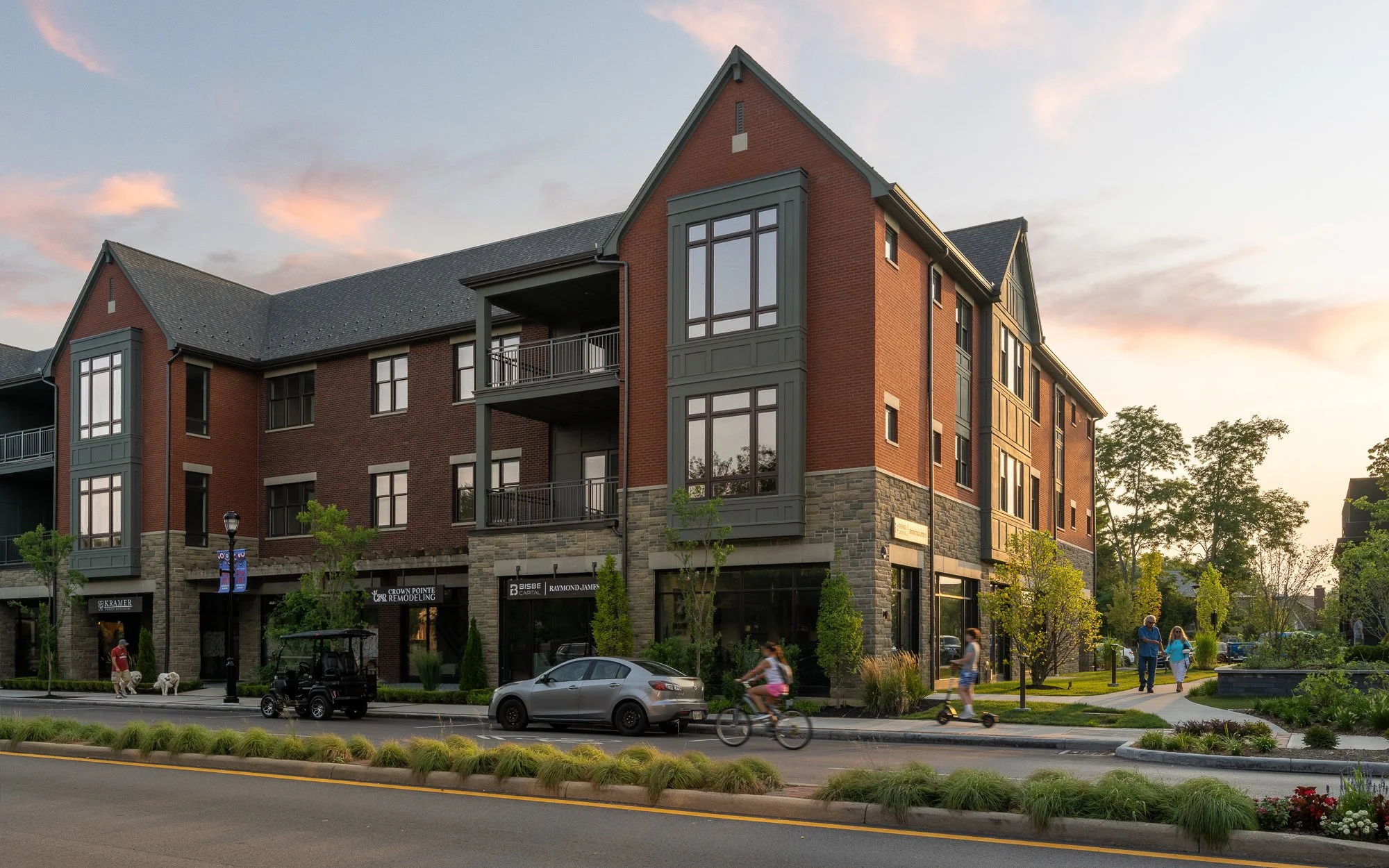 A multi-story brick building with storefronts on the ground level, including a remodeling office and real estate agency, with residential units above. People are walking, biking, and riding scooters along the street, with trees and landscaped greener