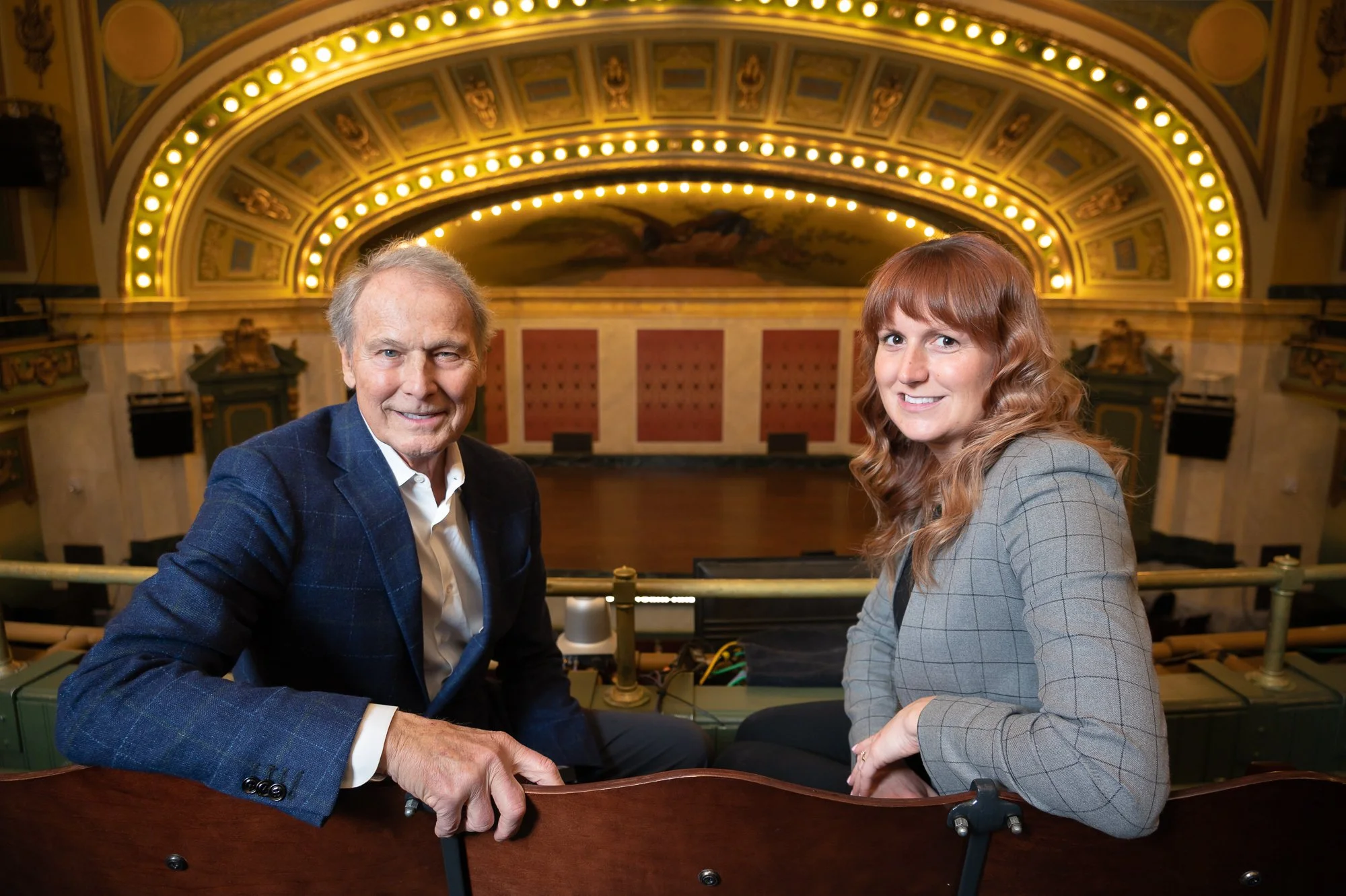 A man and a woman sitting in an ornate theater or opera house with a decorated ceiling and stage in the background.