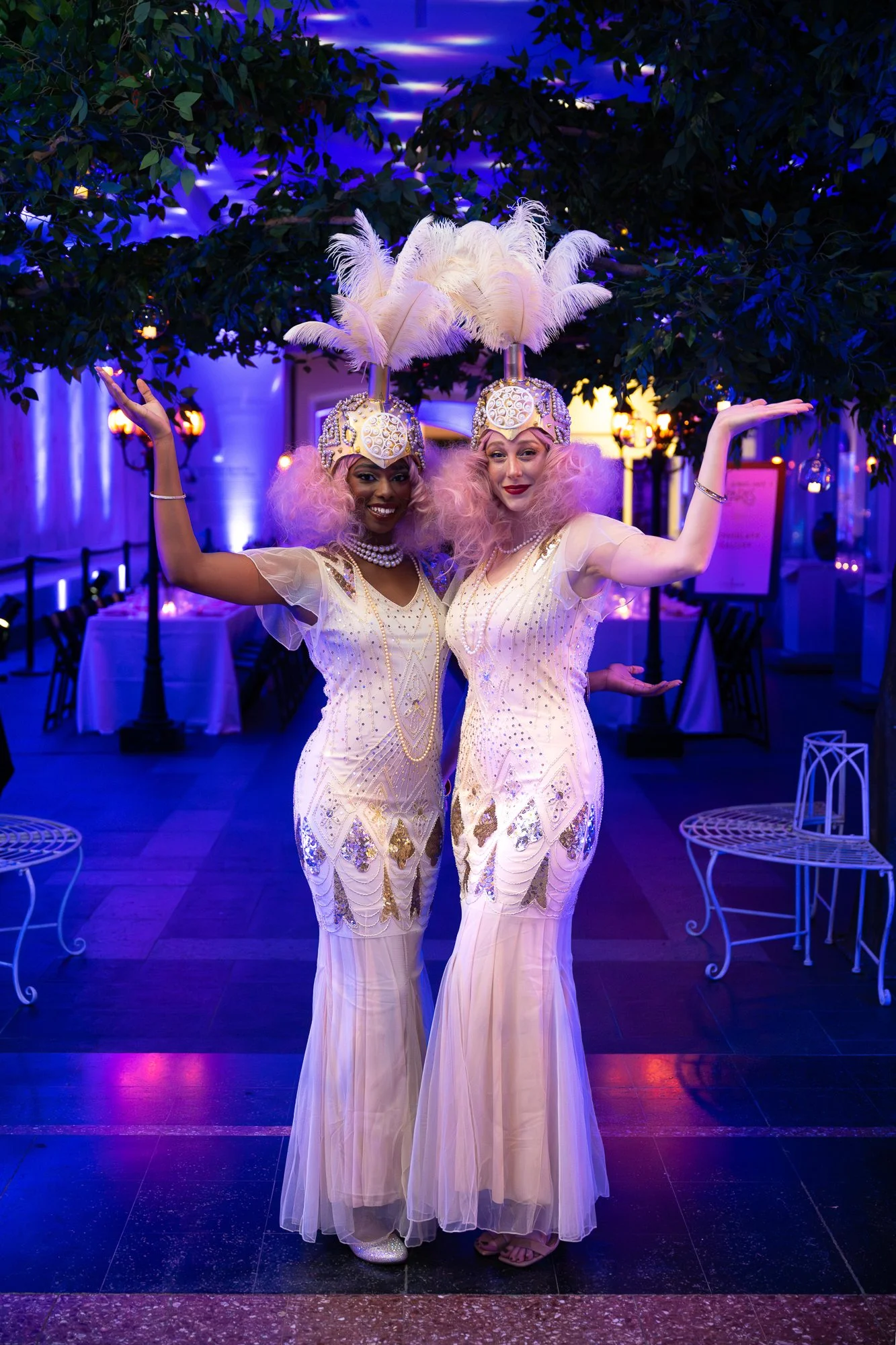 Two women dressed in vintage-style white and gold flapper dresses with feather headpieces, posing happily at an event with purple lighting and decorated indoor setting.