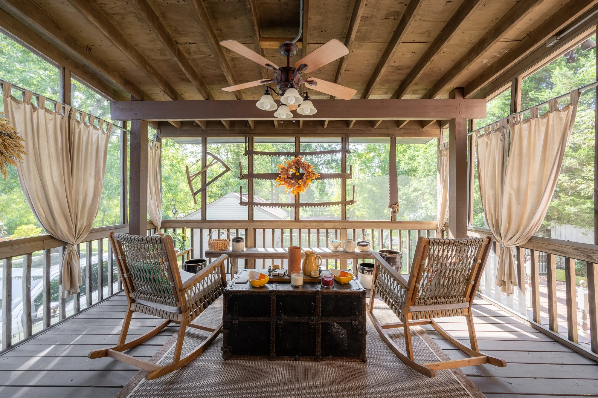 Screened-in porch with wooden ceiling, ceiling fan, two wooden rocking chairs, a black trunk, and a small table with breakfast bowls, pitchers, and other items. Curtains and natural light from large windows with outdoor greenery visible.