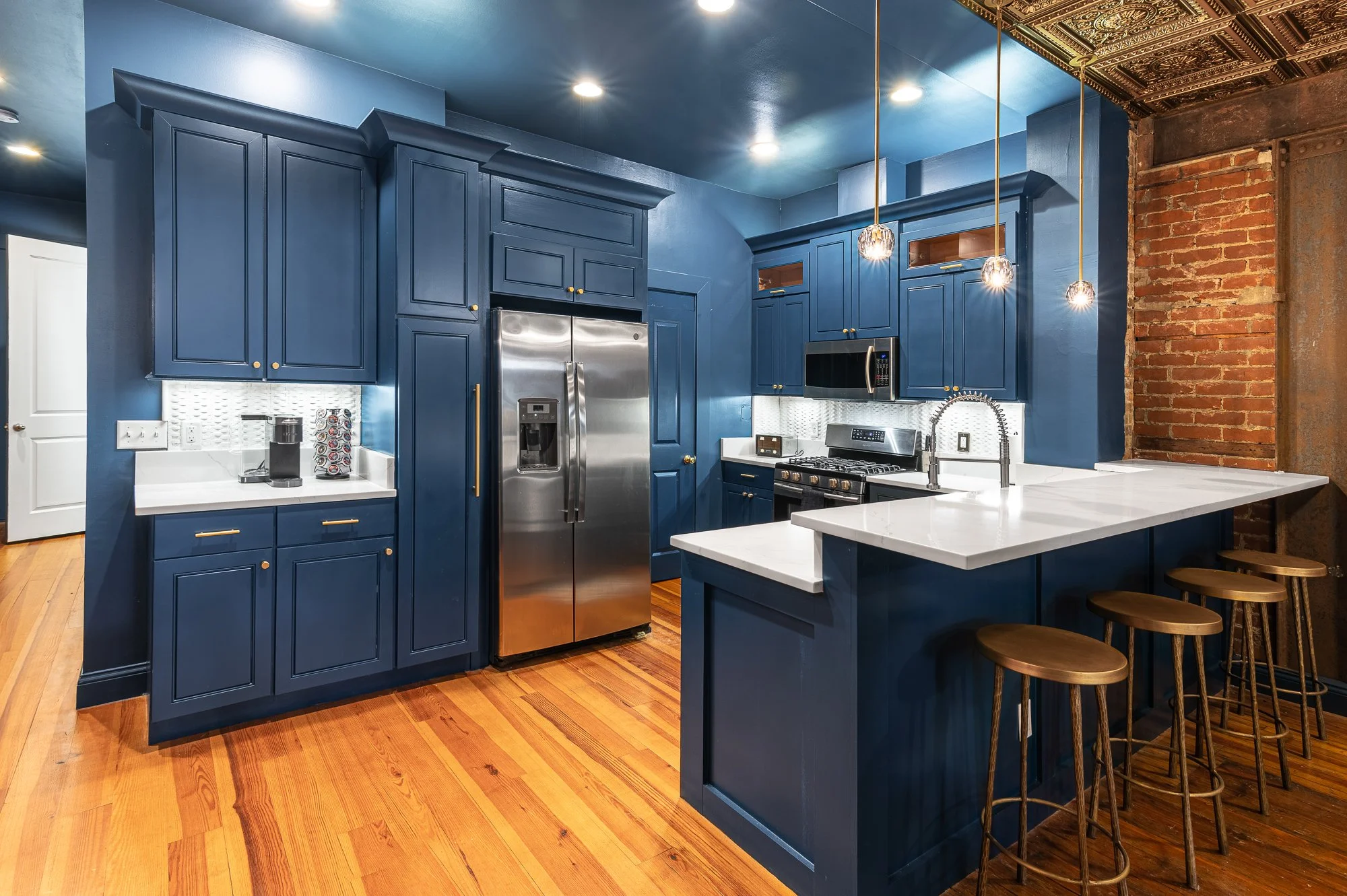 Modern kitchen with blue cabinets, white countertops, stainless steel appliances, wooden stools, and an exposed brick wall.