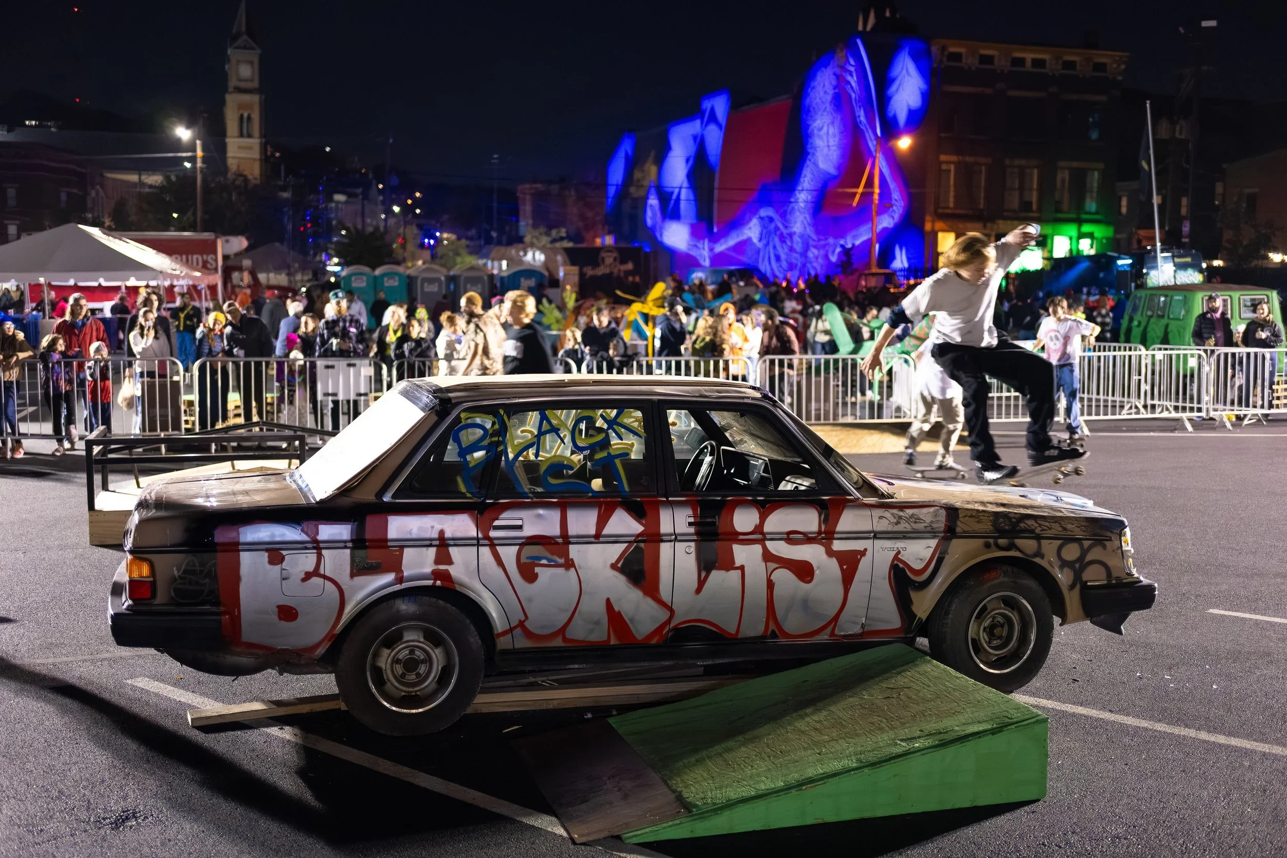 A car with graffiti on it, parked on a ramp at night during a street festival, with a crowd and amusement rides in the background.