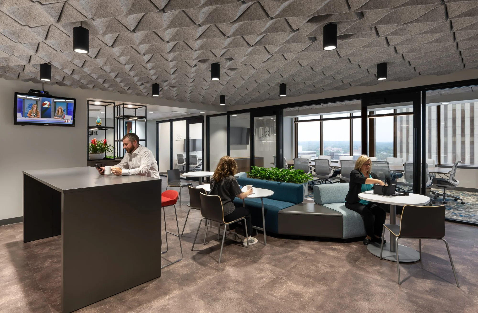 Modern office lounge with three people, two women and one man, working and relaxing. The space features a textured ceiling, large windows with city views, and various seating arrangements, including chairs, a curved sofa, and a high counter. A TV dis