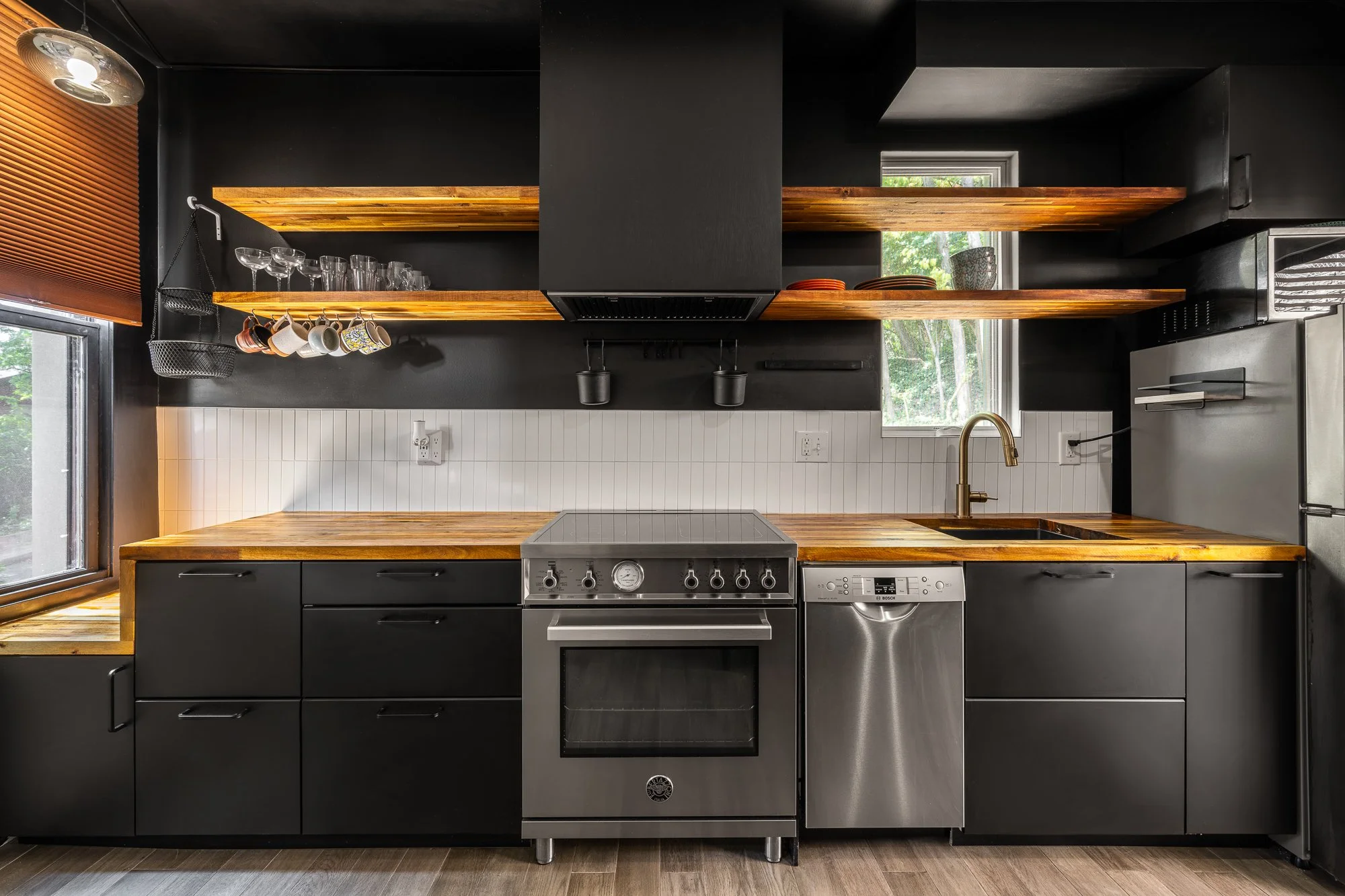 Modern kitchen with black cabinets, wooden countertops and open wooden shelves, stainless steel oven and dishwasher, window with a view of greenery, and black wall with hanging cups and glasses.