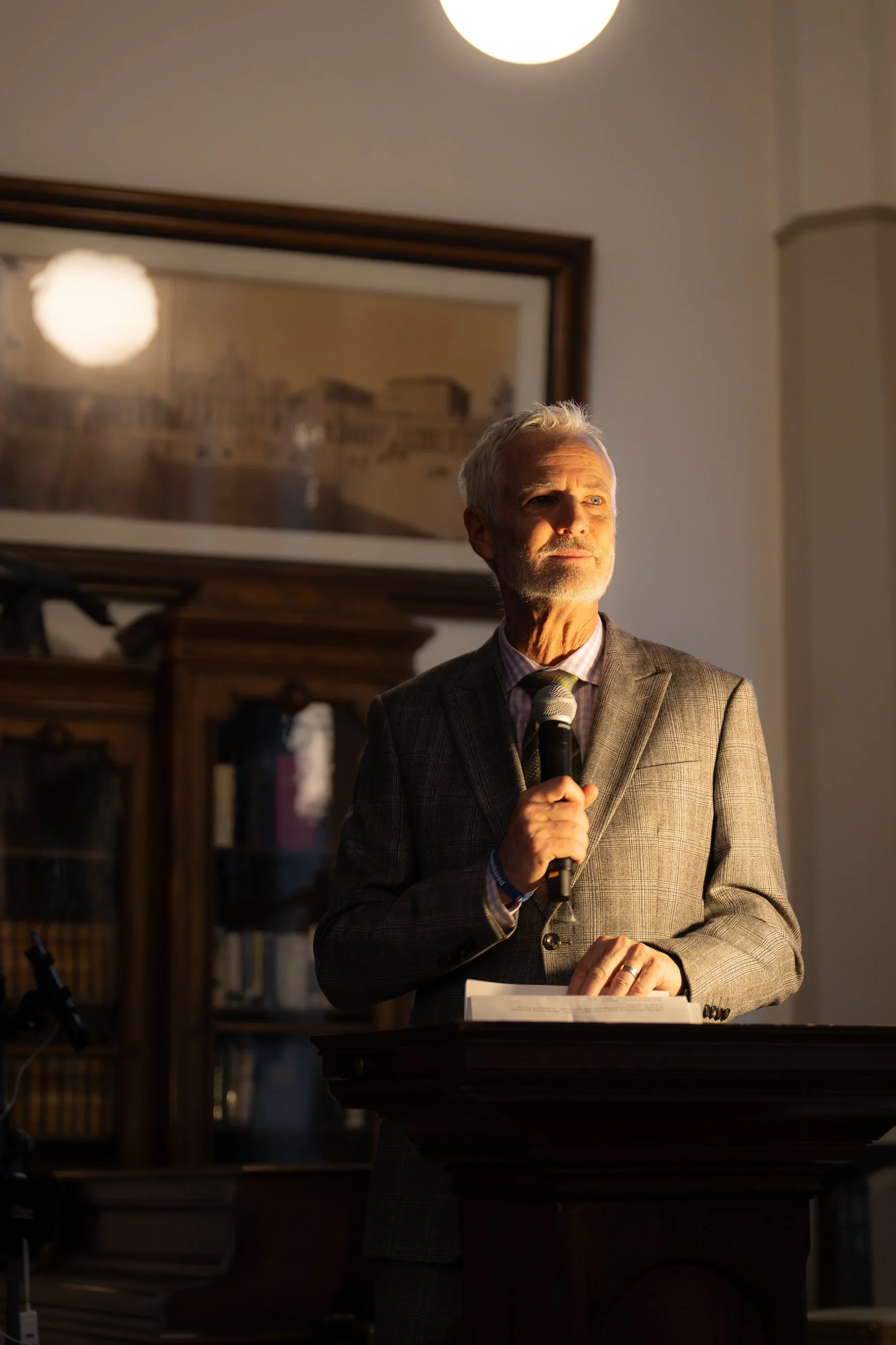An elderly man with gray hair and a beard, dressed in a gray plaid suit, holding a microphone and a folder, speaking at a formal event in a room with wooden furniture and framed pictures on the wall.