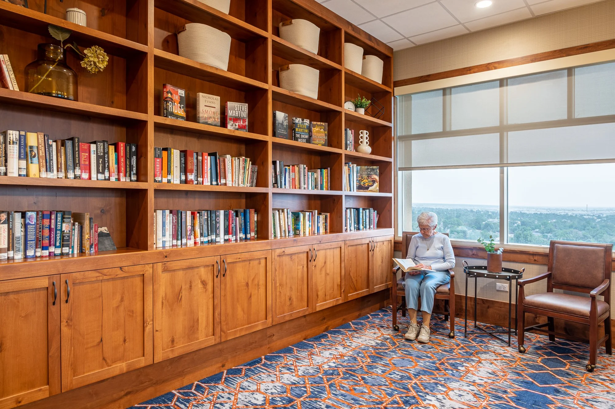 An elderly woman sitting in a cozy room near a large window, reading a book. The room features a tall wooden bookshelf filled with books and decorative items, a small side table with a potted plant, and a patterned rug on the floor.
