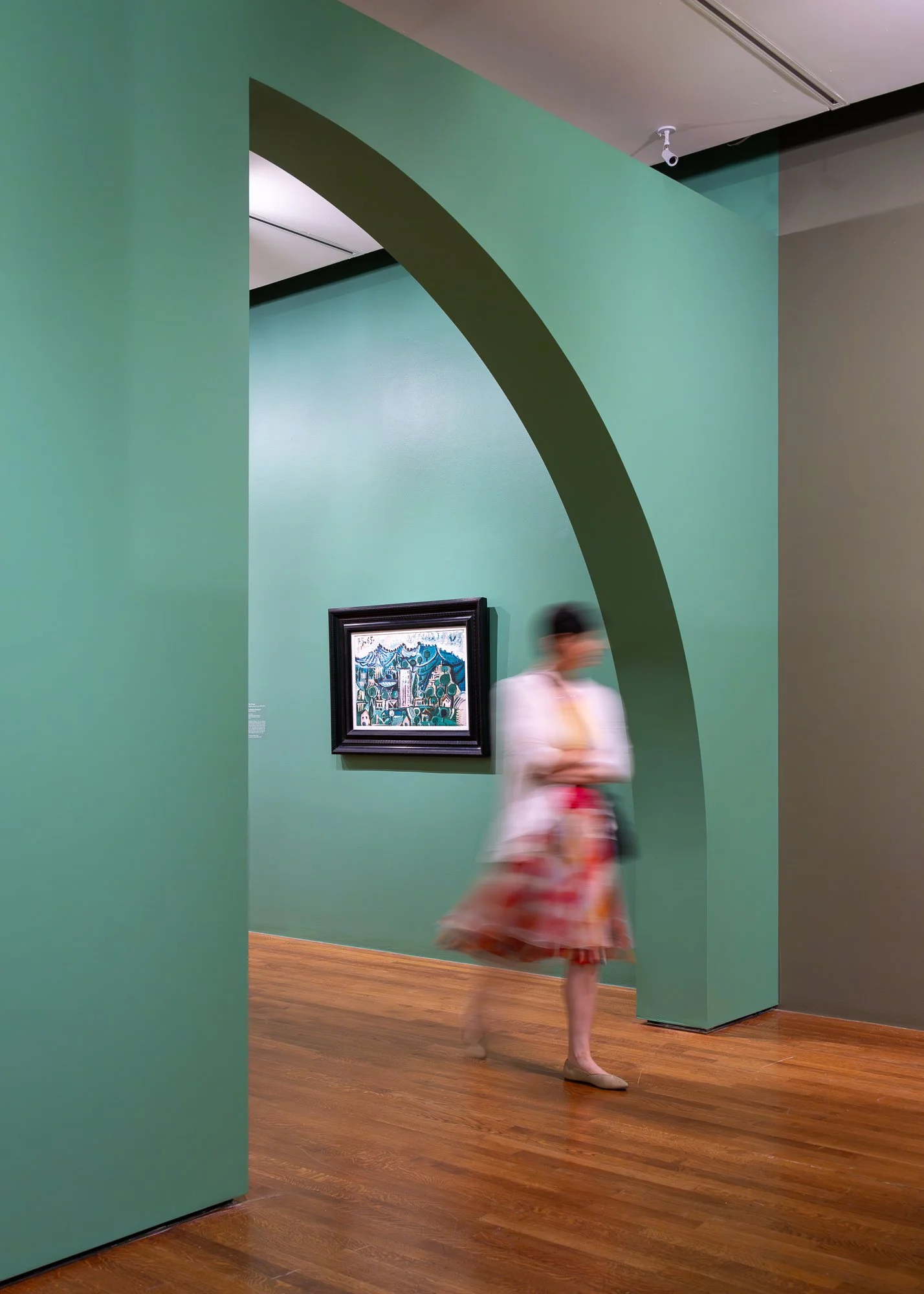 A blurred woman in a floral skirt and beige shoes walking past a framed artwork on a green wall inside an art gallery.