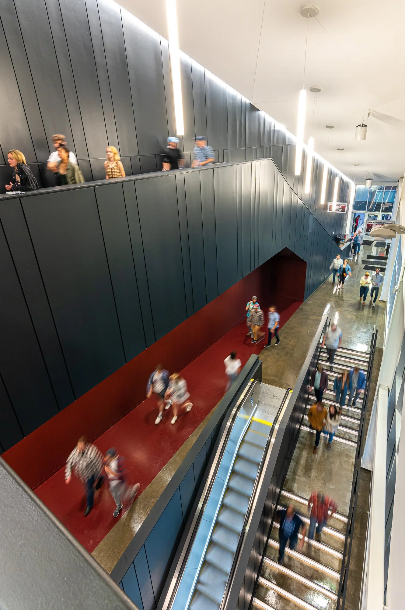 People walking and descending escalators in a modern indoor public space, possibly an airport.